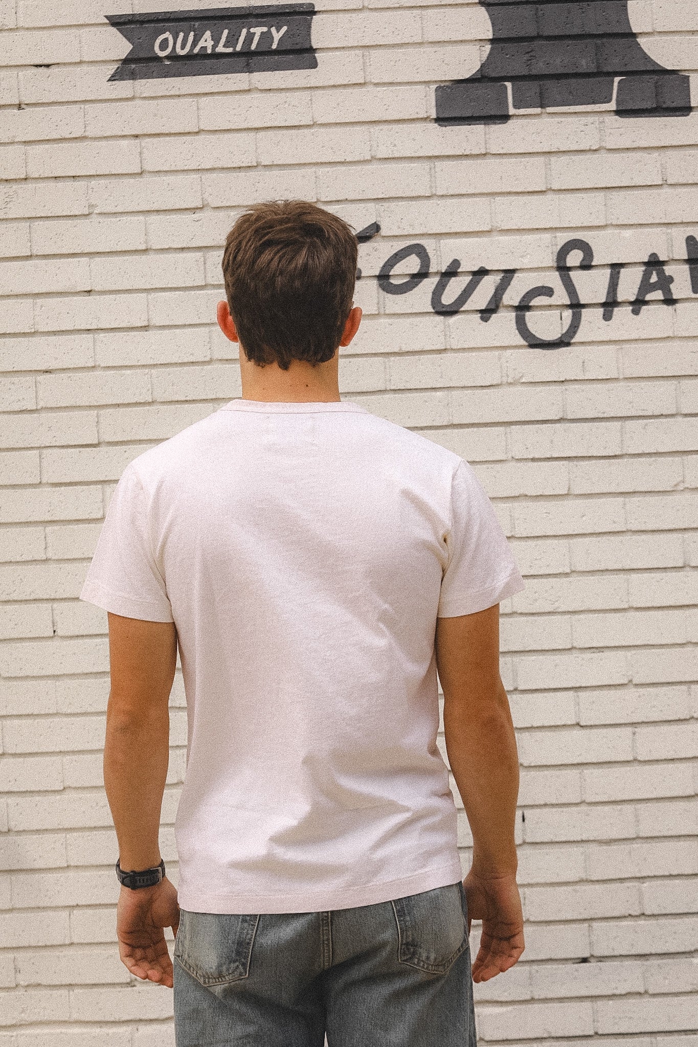 Man wearing a white t-shirt standing in front of a white brick wall with graffiti.