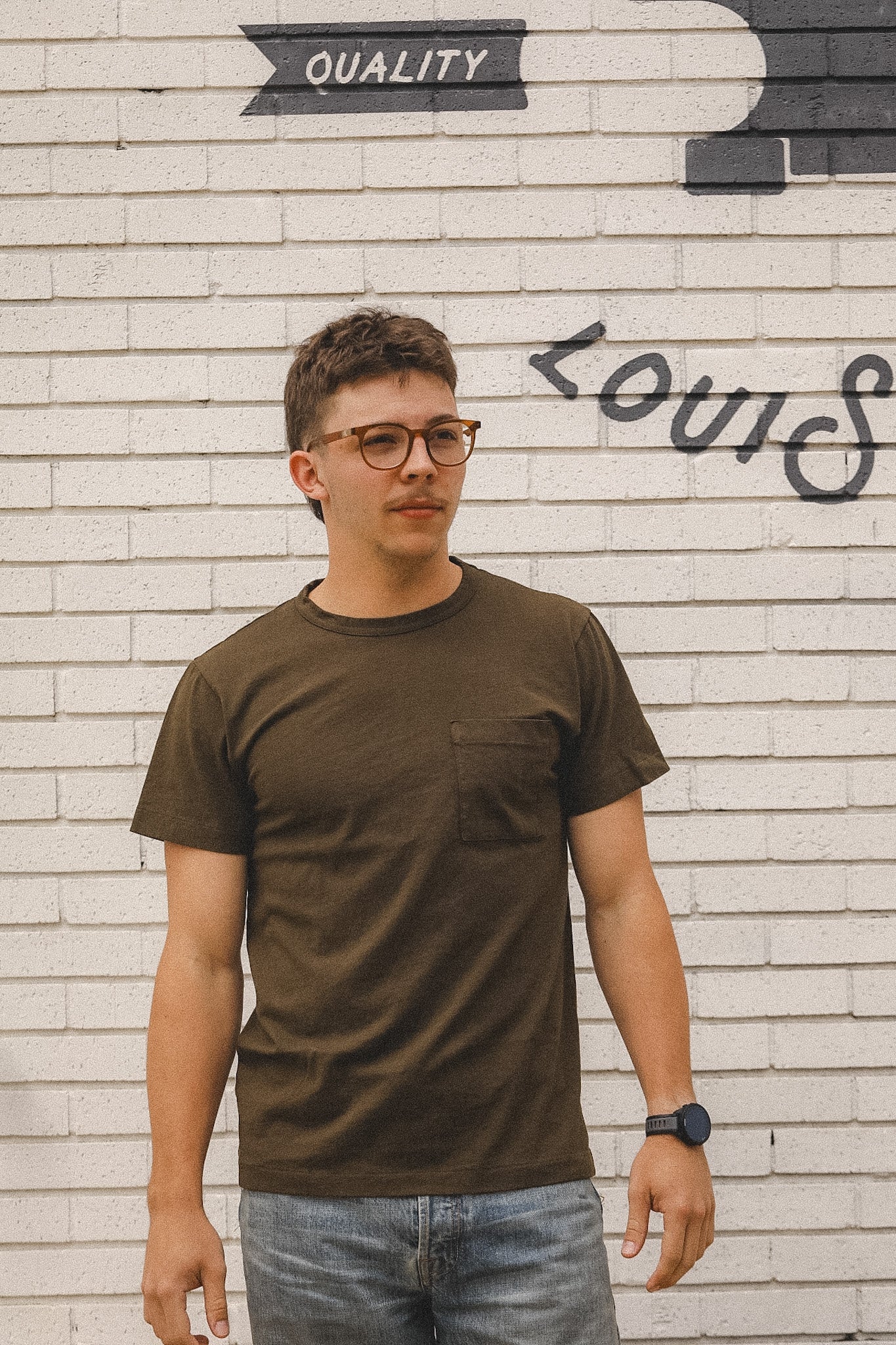 Man wearing a brown t-shirt and glasses standing in front of a brick wall with graffiti.
