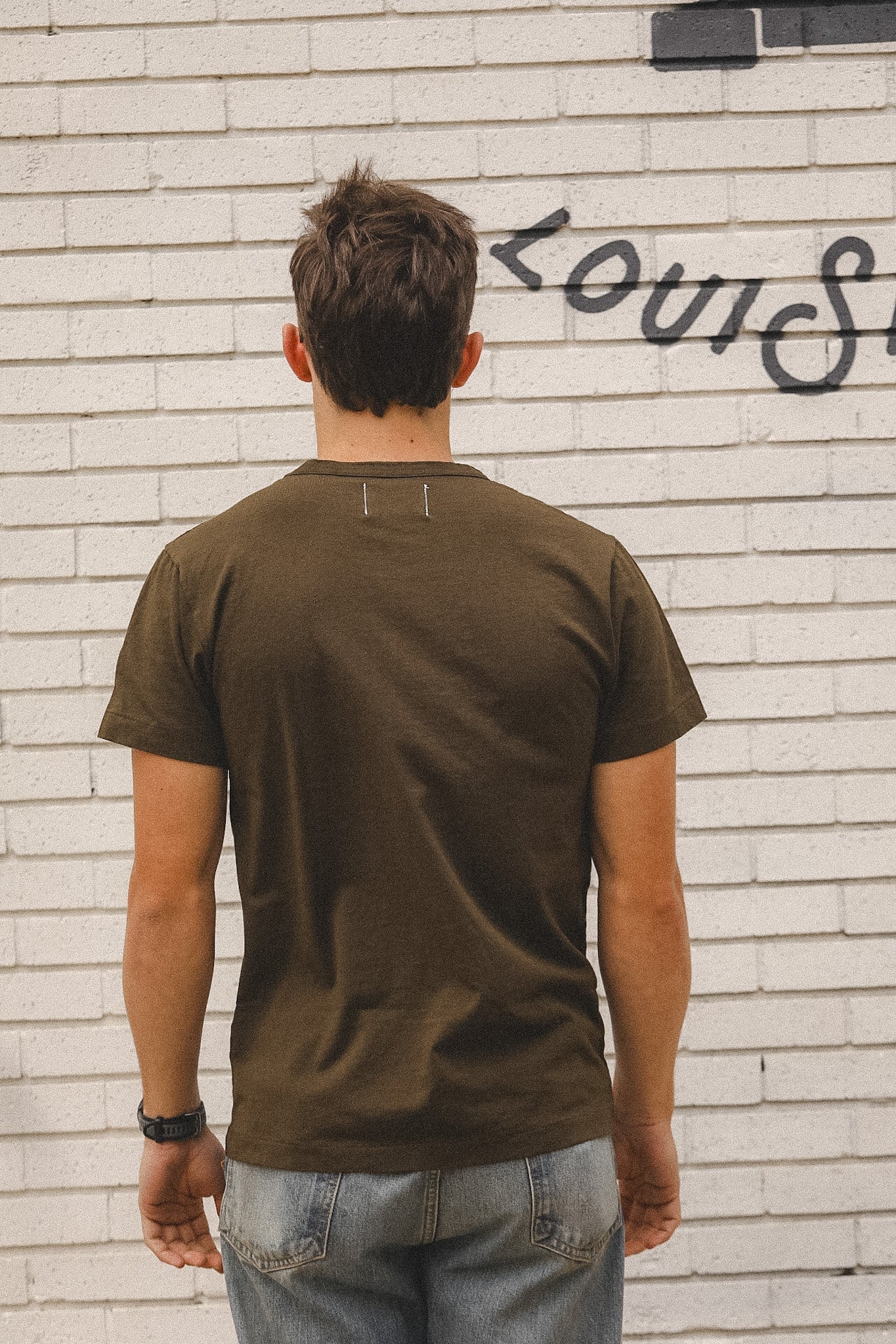 Man wearing a brown t-shirt standing in front of a white brick wall with graffiti.