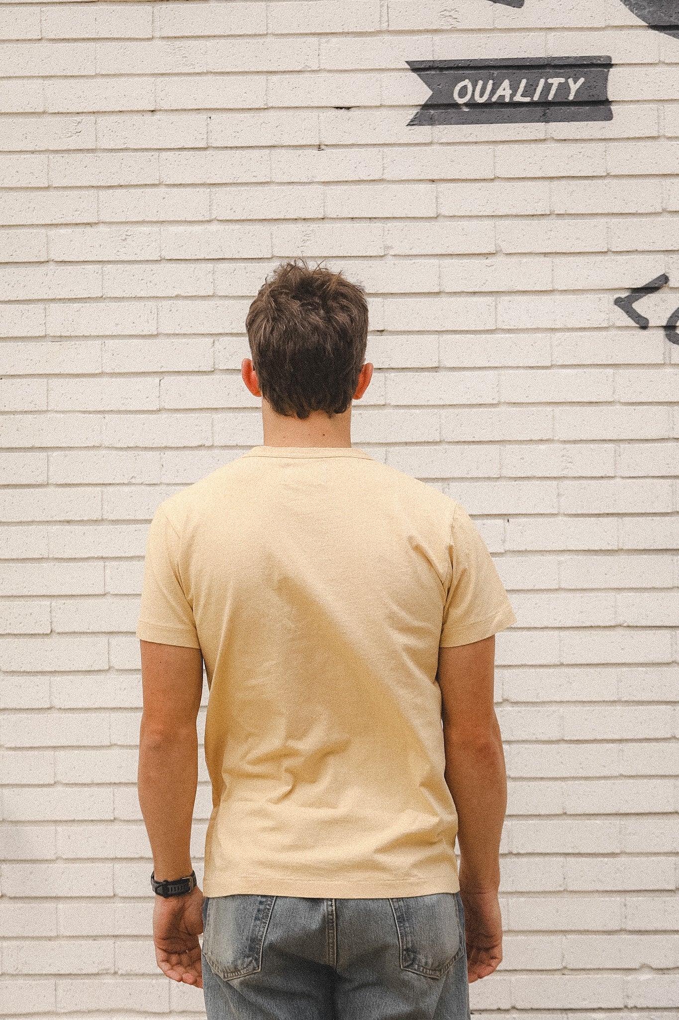 Man wearing a beige t-shirt standing in front of a textured wall with 'QUALITY' text.
