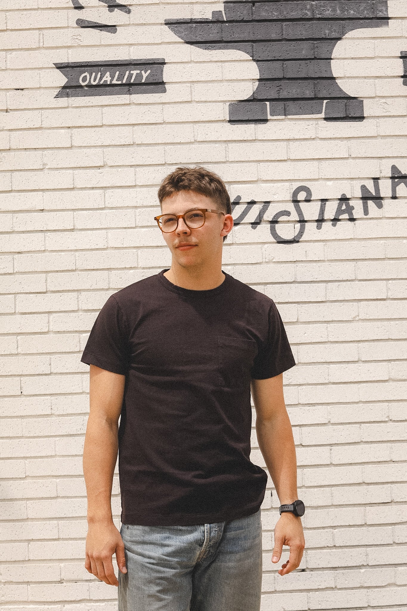 Person wearing a black t-shirt and glasses standing in front of a brick wall with graffiti.
