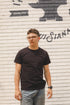 Person wearing a black t-shirt and glasses standing in front of a brick wall with graffiti.