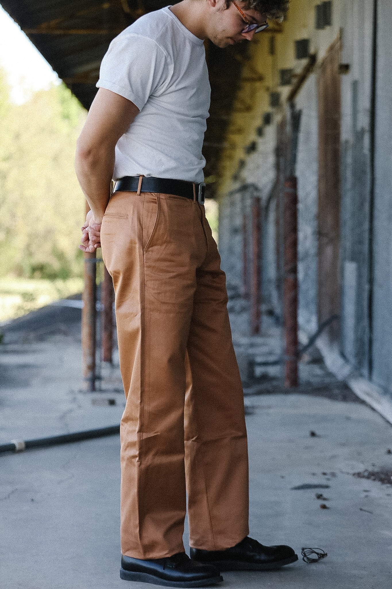 Man wearing a white t-shirt and brown pants standing on a concrete surface with a building in the background.