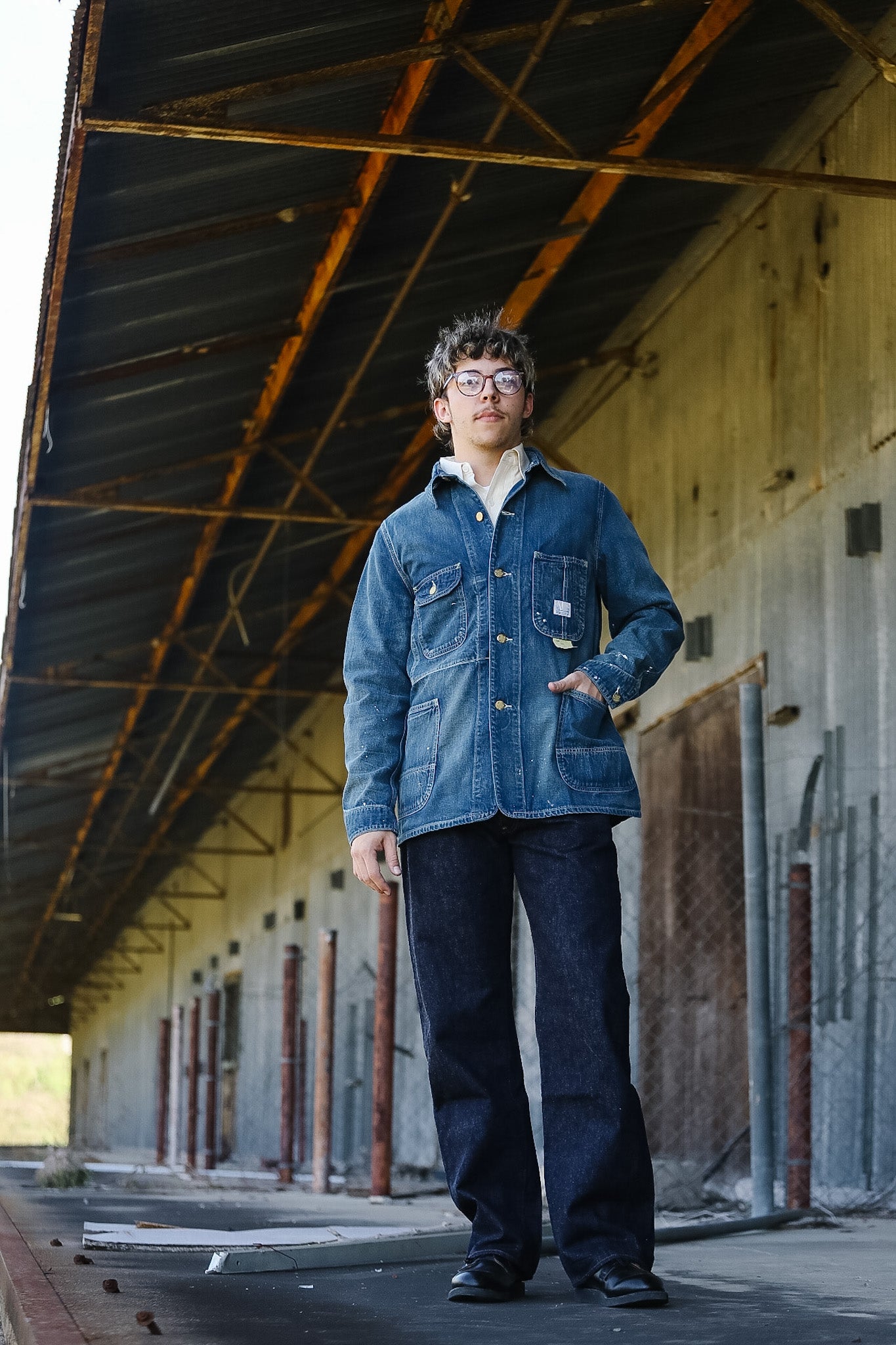 Person wearing a blue denim jacket and dark pants standing in an industrial setting with rusted metal beams.