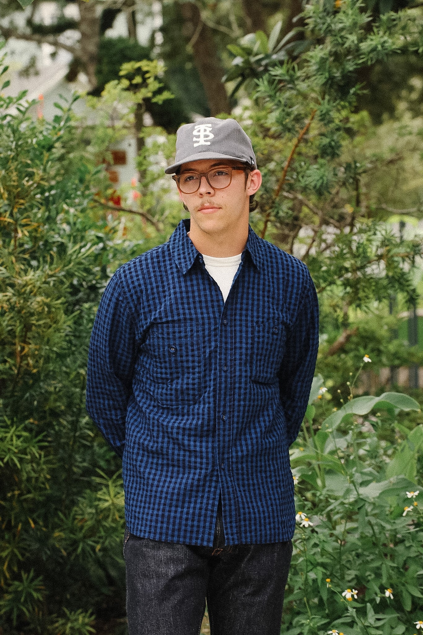 A man wearing a blue and black checkered button-up shirt, dark jeans, a gray baseball cap with a white logo, and glasses, standing outdoors in front of green foliage.