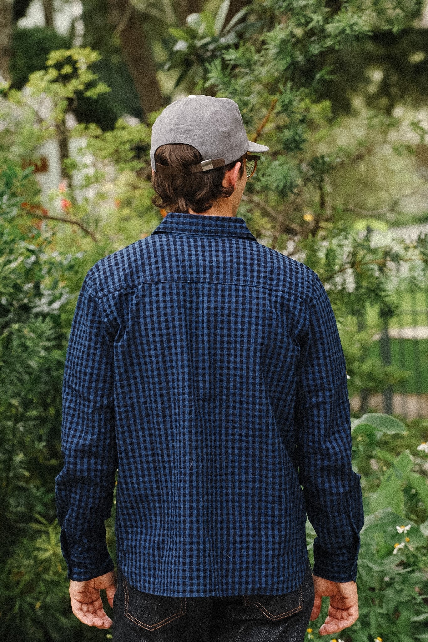 A man viewed from behind wearing a dark blue and black checkered long-sleeve shirt, dark jeans, and a gray baseball cap, standing outdoors in a garden setting.