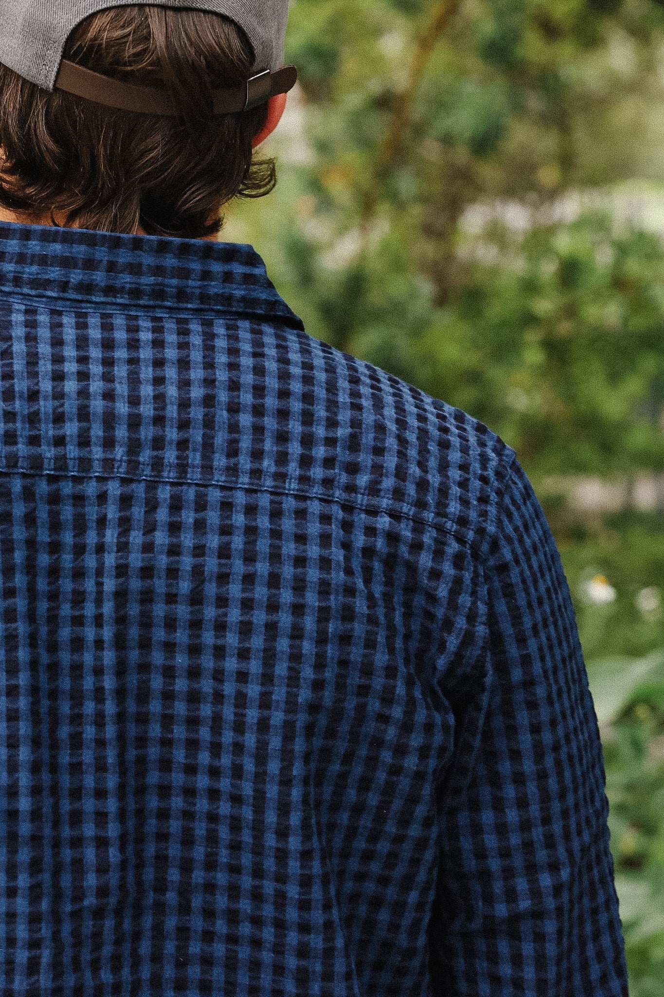 A person viewed from behind wearing a dark blue and black gingham plaid button-up shirt, with a gray cap, against a blurred green outdoor background.