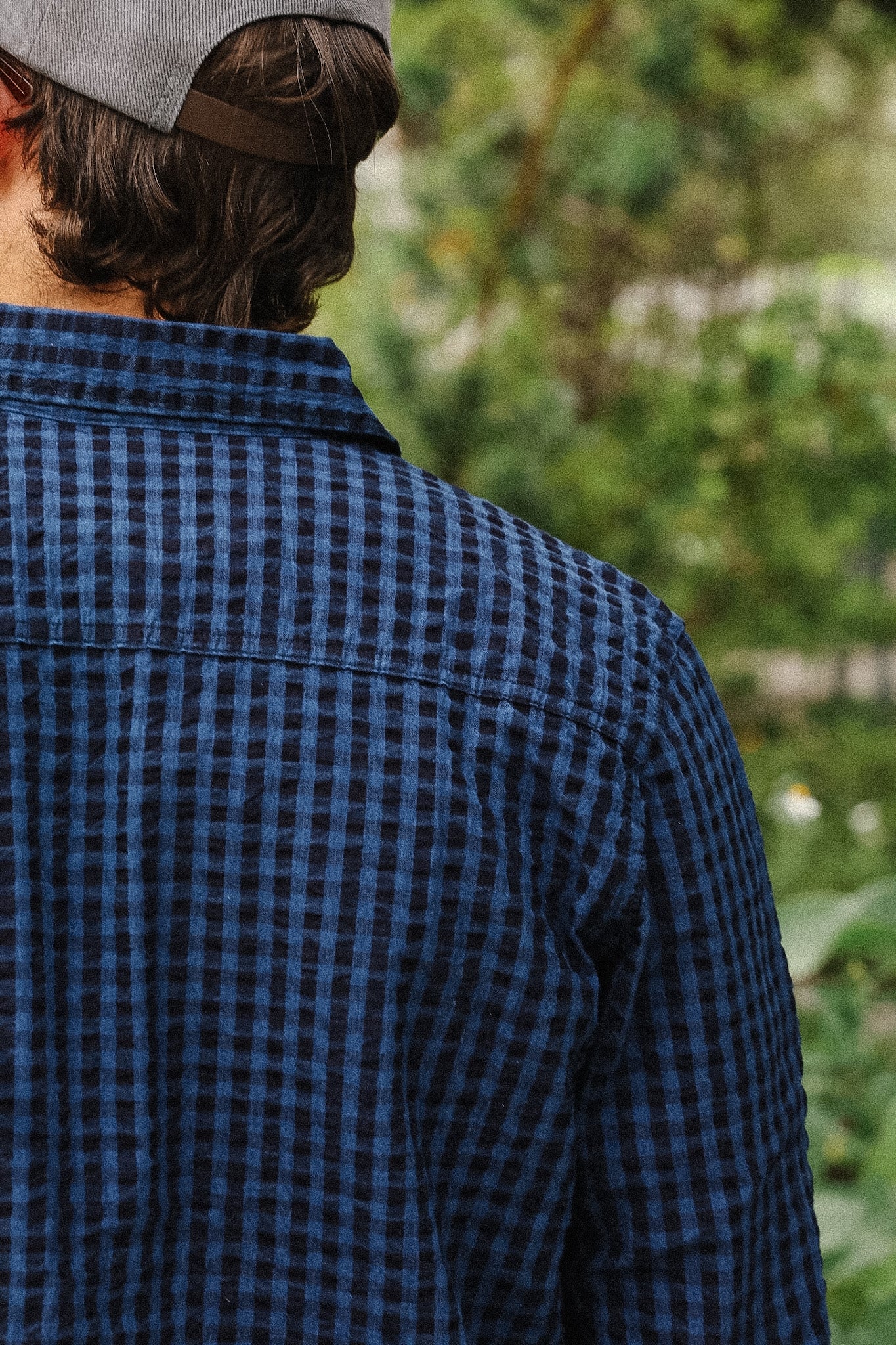 A person viewed from behind wearing a dark blue and black checkered flannel shirt and a gray cap, with a blurred green outdoor background.