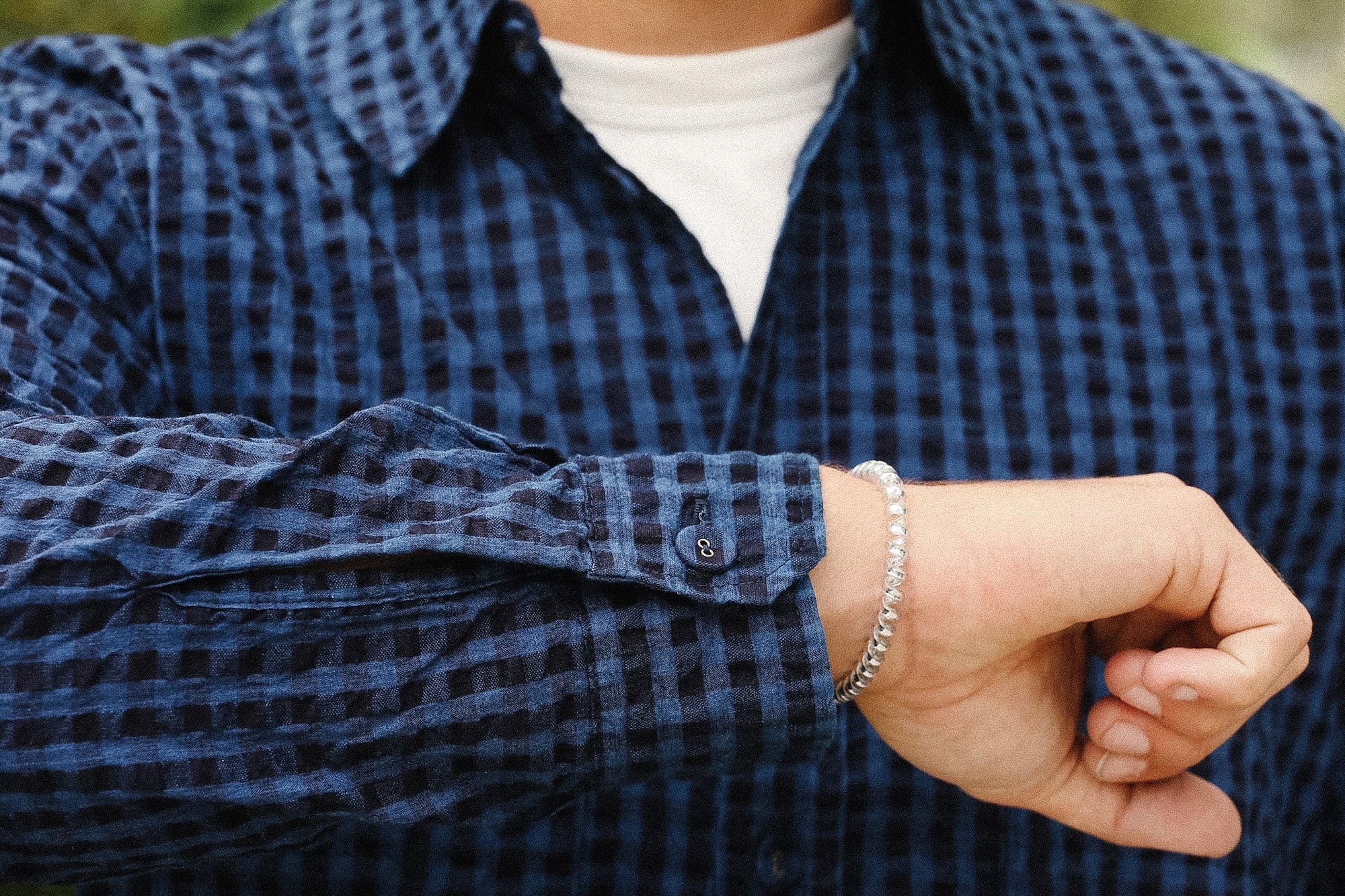 A close-up of a person's wrist wearing a silver chain bracelet, wearing a blue and black checkered flannel shirt over a white t-shirt.