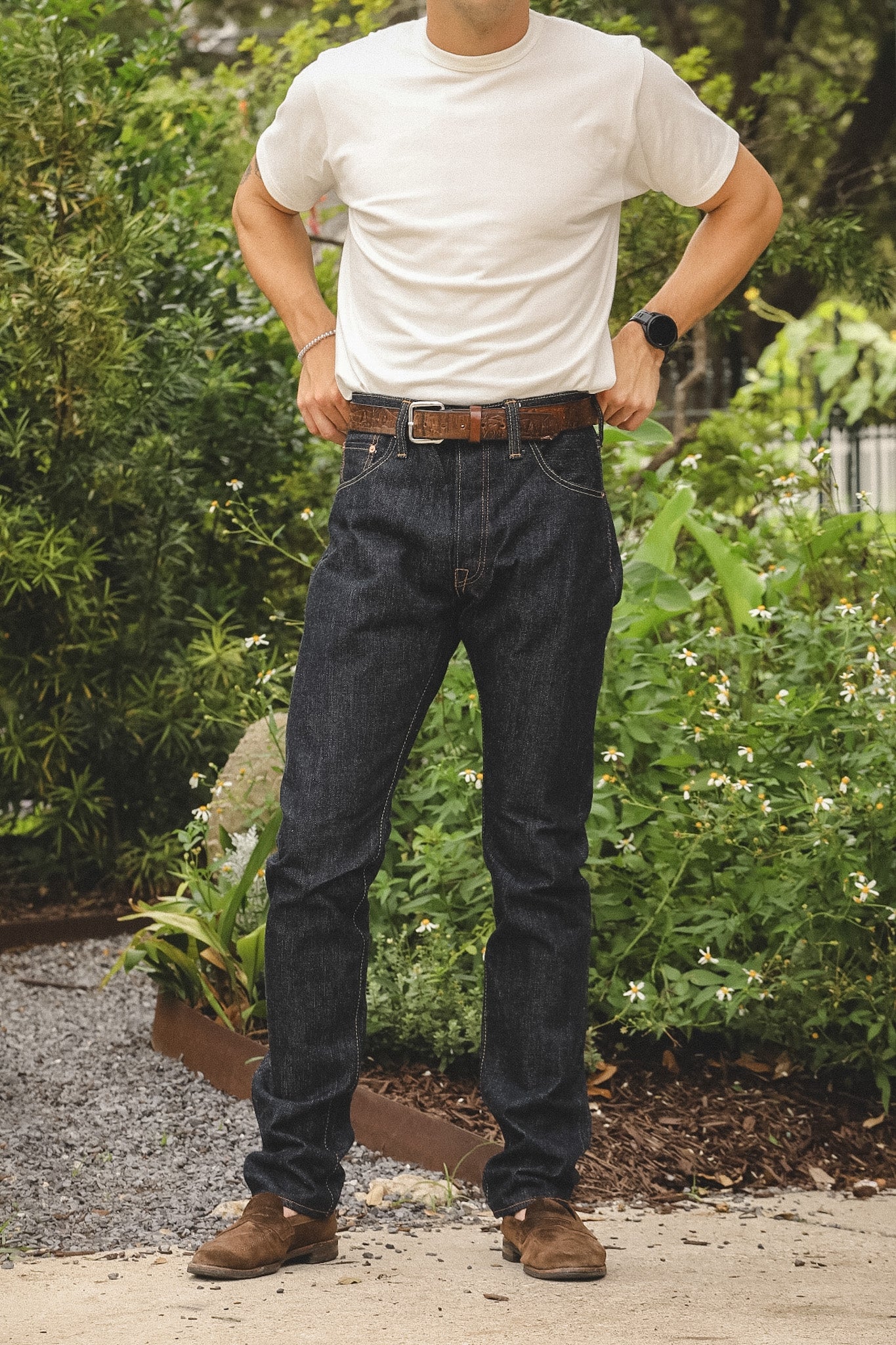 A man wearing dark wash blue jeans, a white t-shirt, and brown leather shoes, standing outdoors in front of green garden foliage.