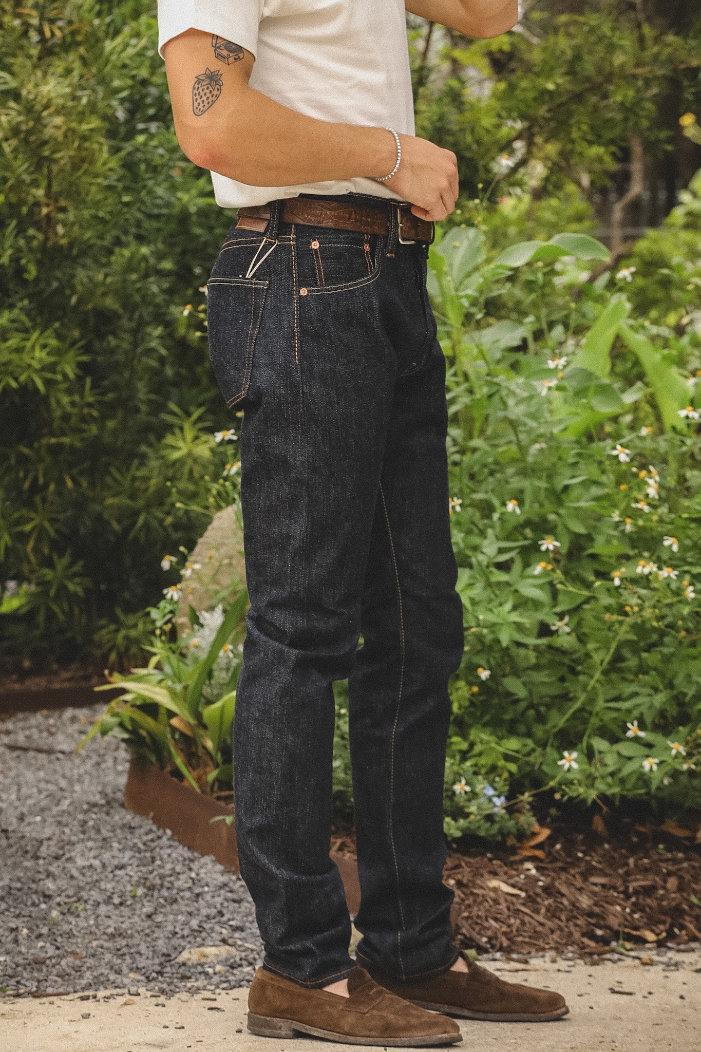 A person wearing dark indigo denim jeans, a white t-shirt, and brown suede shoes, standing outdoors in a garden setting.