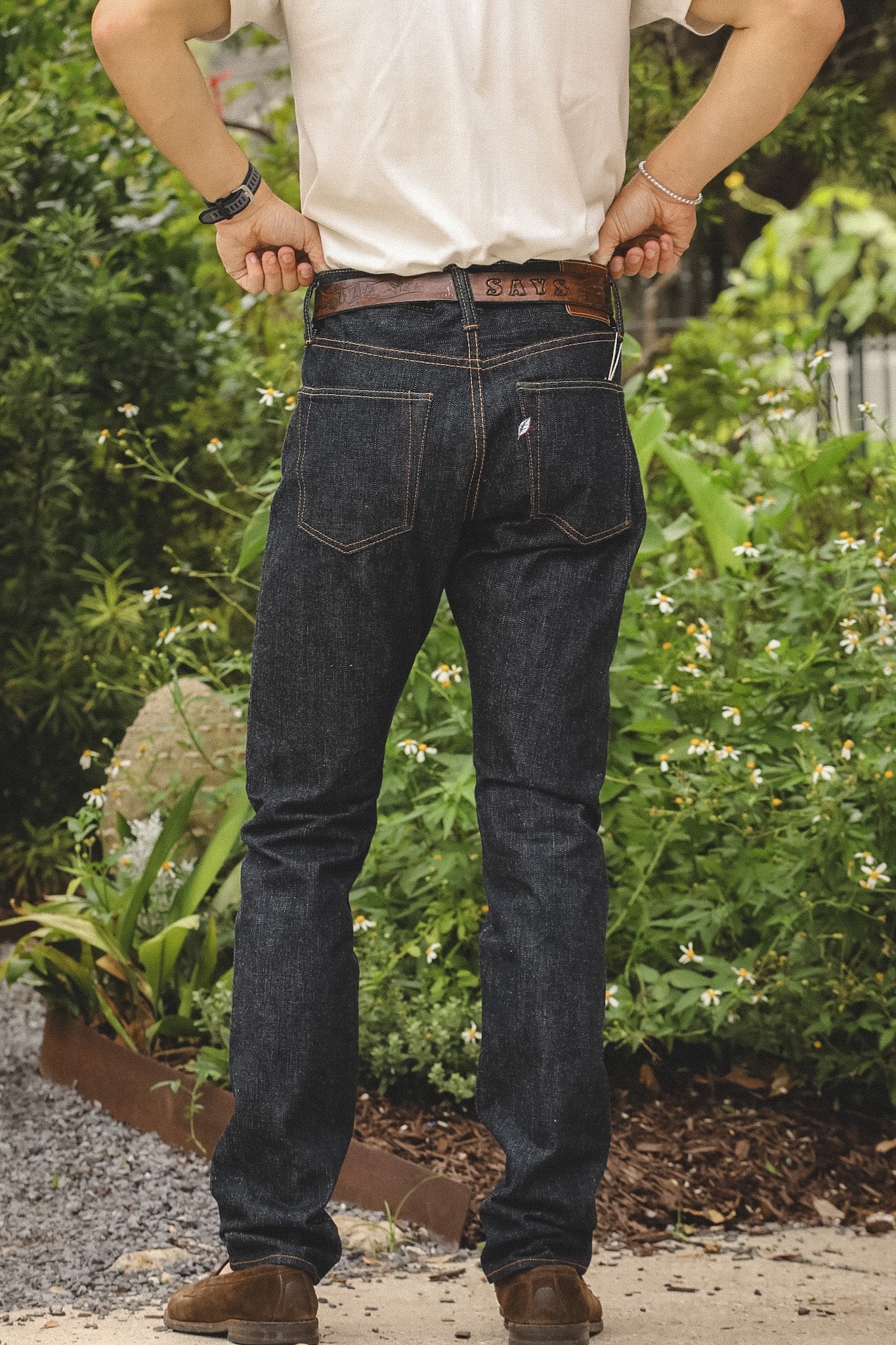 A man viewed from behind wearing dark indigo denim jeans, a white t-shirt, and brown boots, standing outdoors in a garden setting.