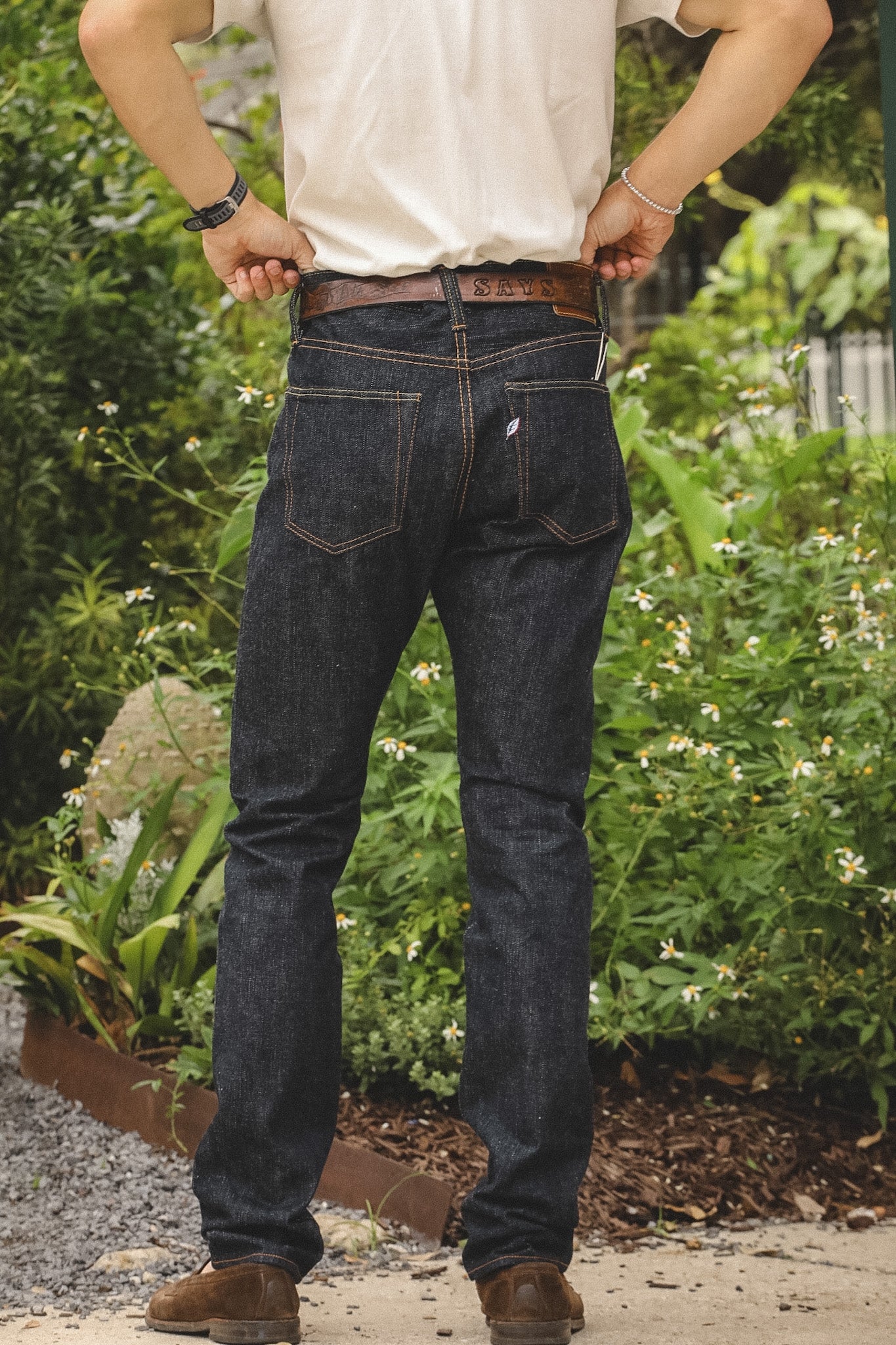 A person viewed from the back wearing dark indigo jeans, a white t-shirt, and brown boots, standing outdoors in front of green garden foliage.