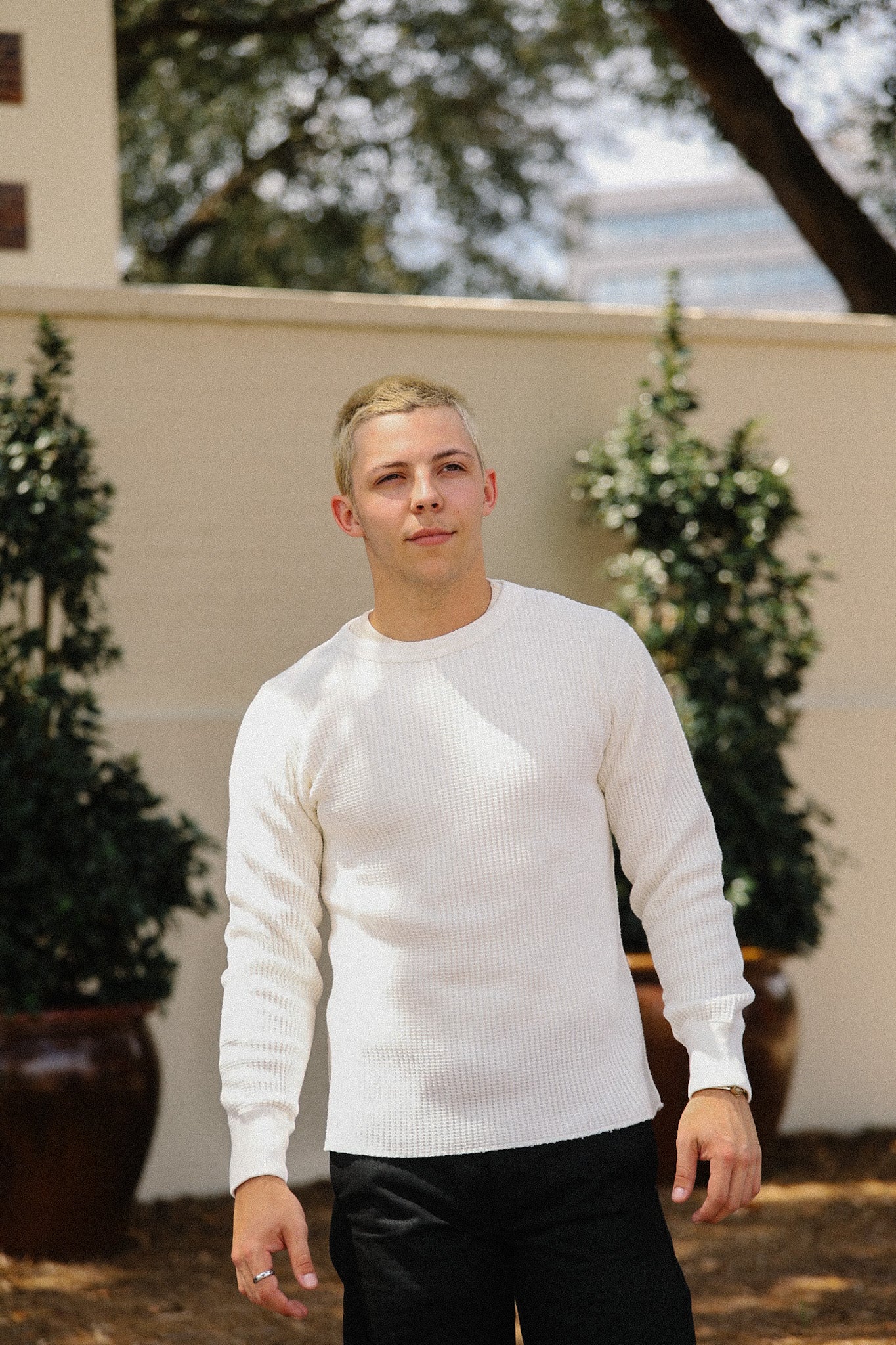 Person wearing a white sweater and black pants standing outdoors with plants in the background