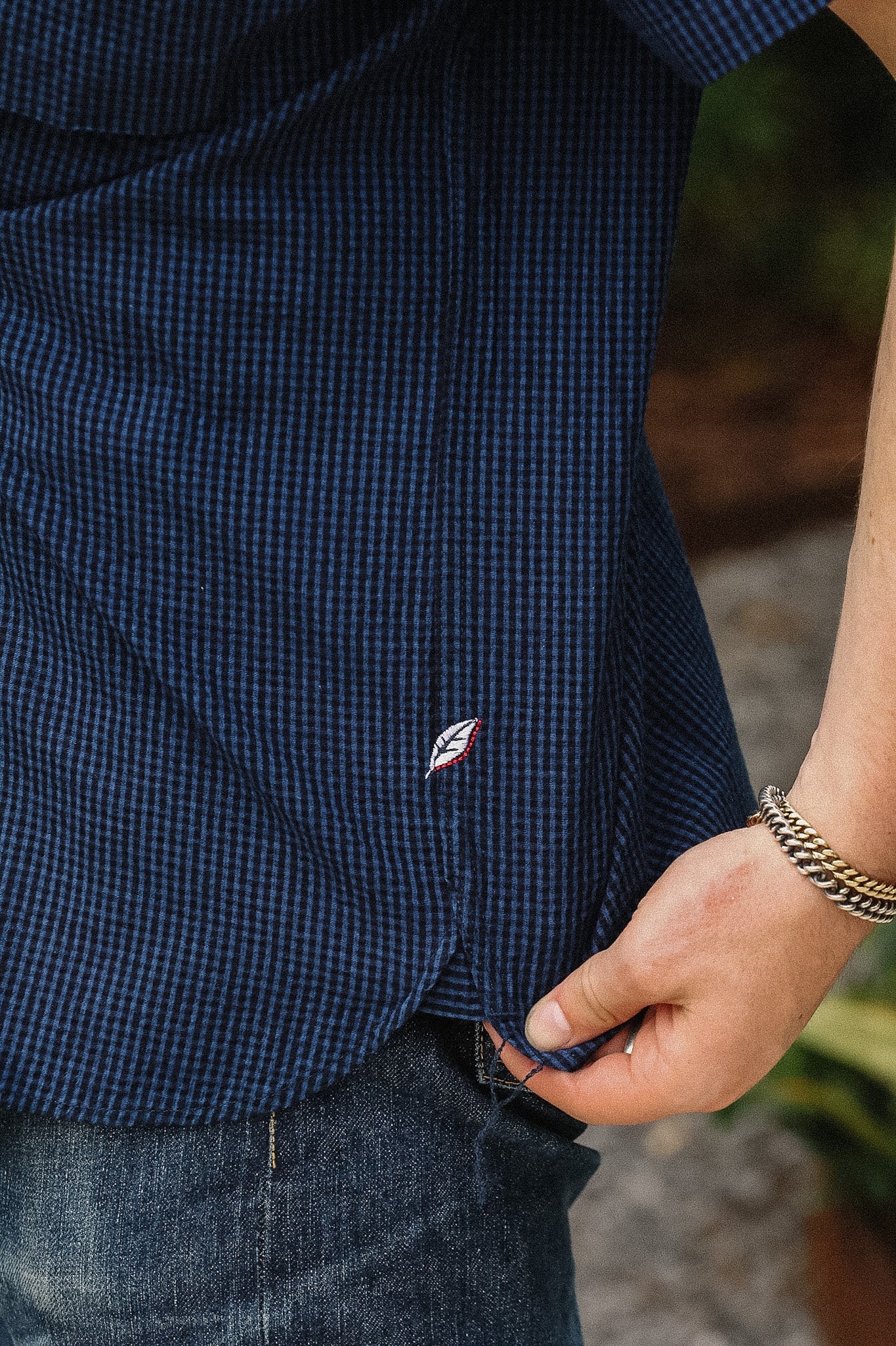 Close-up of a person wearing a navy blue gingham check short-sleeve button-down shirt, pulling the hem slightly with a hand. A small embroidered logo is visible on the lower front, and the person is also wearing blue jeans and a chain bracelet.