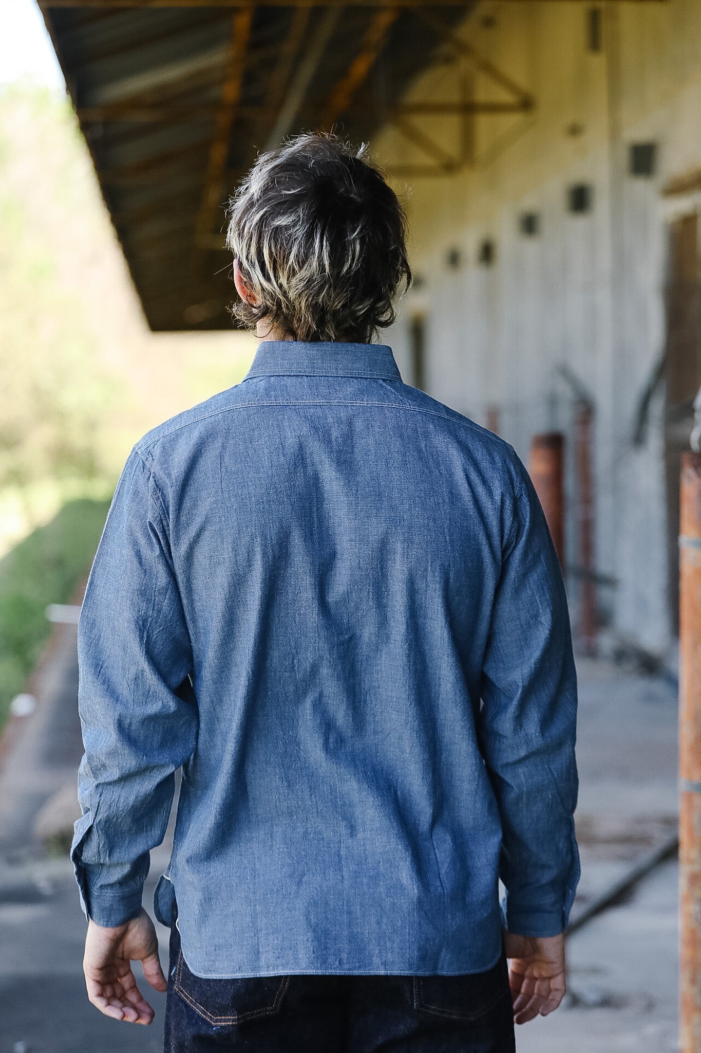 Person wearing a blue denim shirt standing in an outdoor setting with a building in the background.