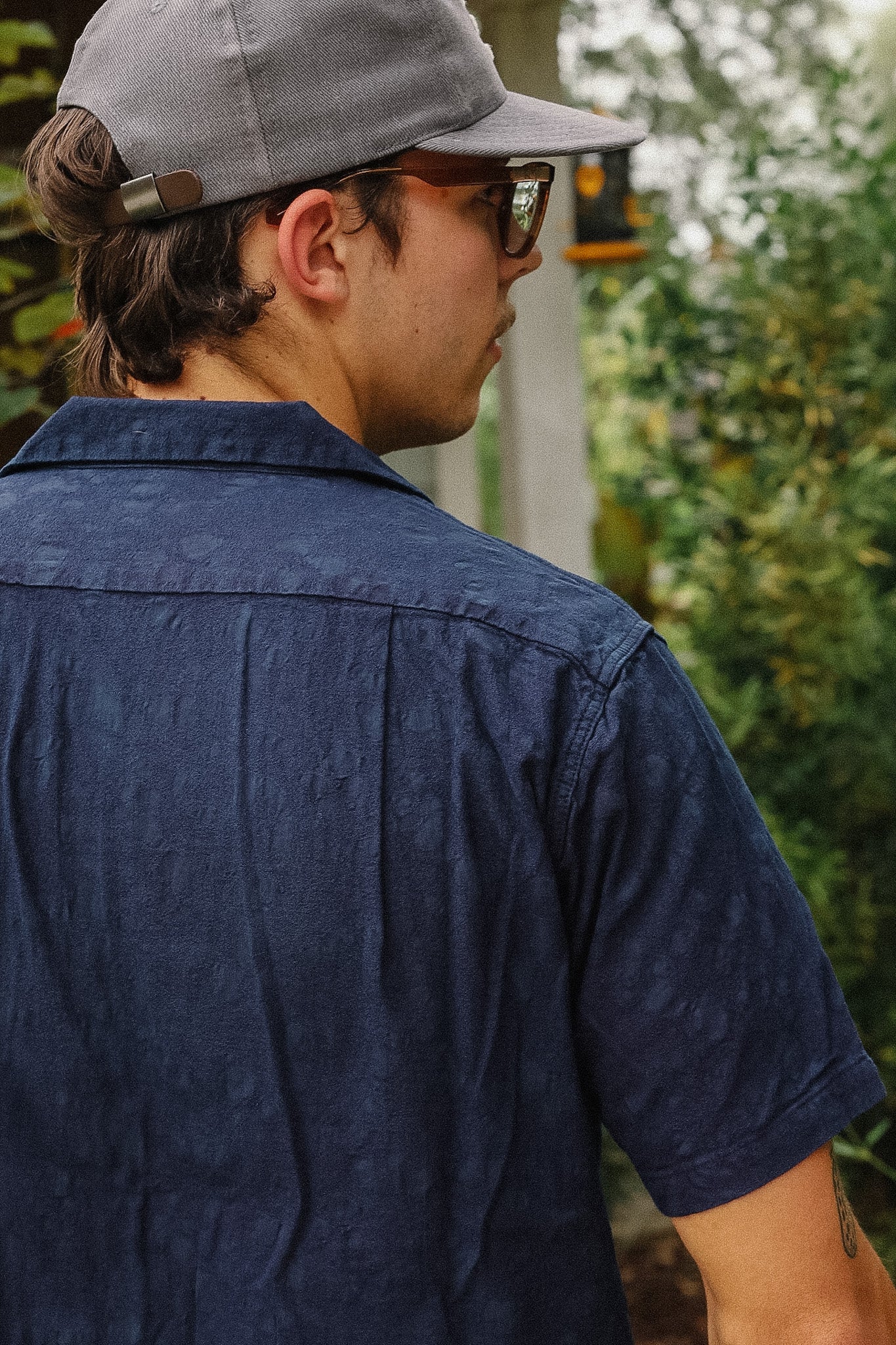 A man wearing a dark navy blue short-sleeve button-up shirt, a gray baseball cap, and glasses, shown from a side-back angle against a blurred green outdoor background.
