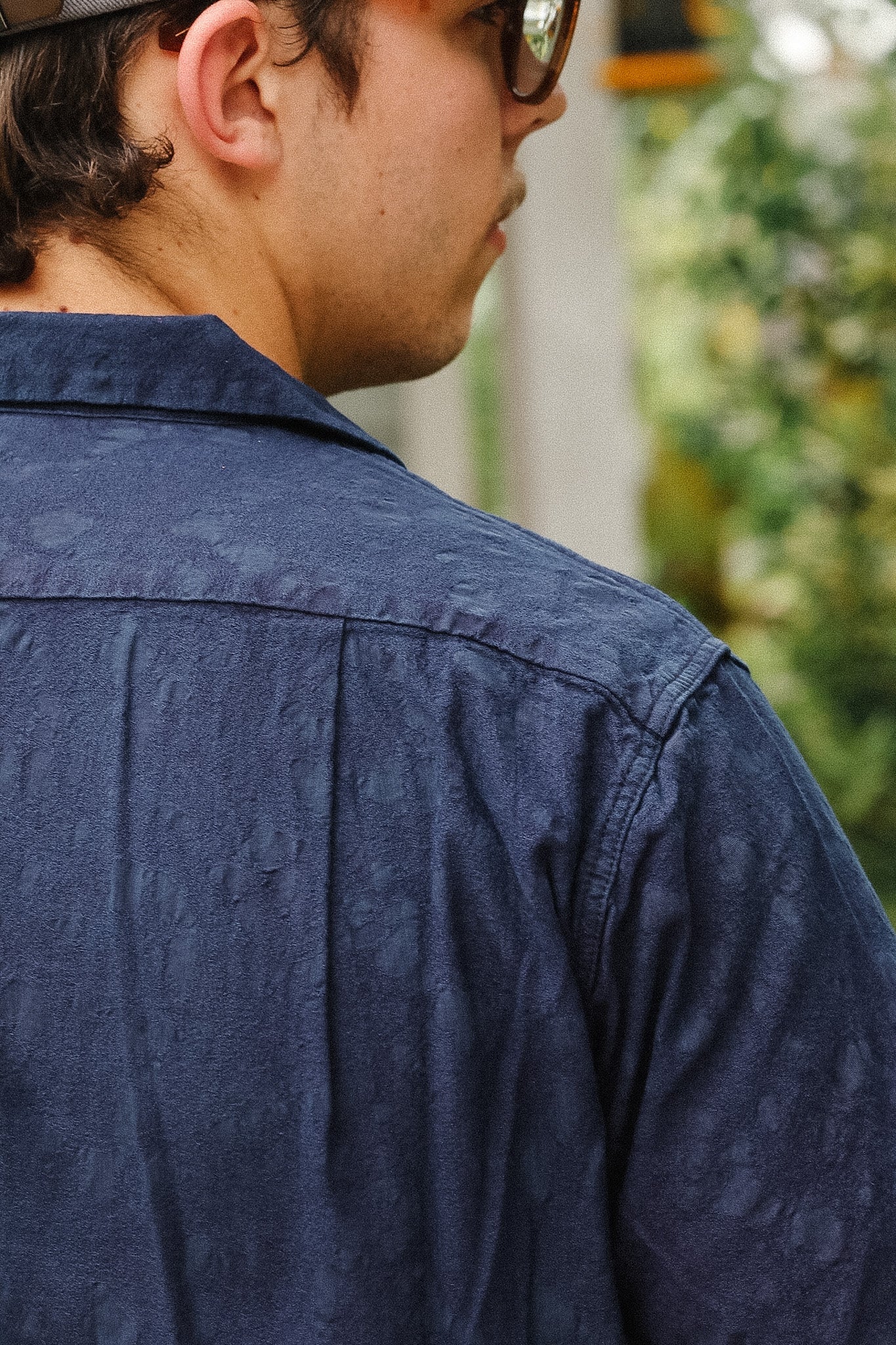 A close-up side profile of a man wearing a textured navy blue button-down shirt, with a blurred outdoor background of green foliage.