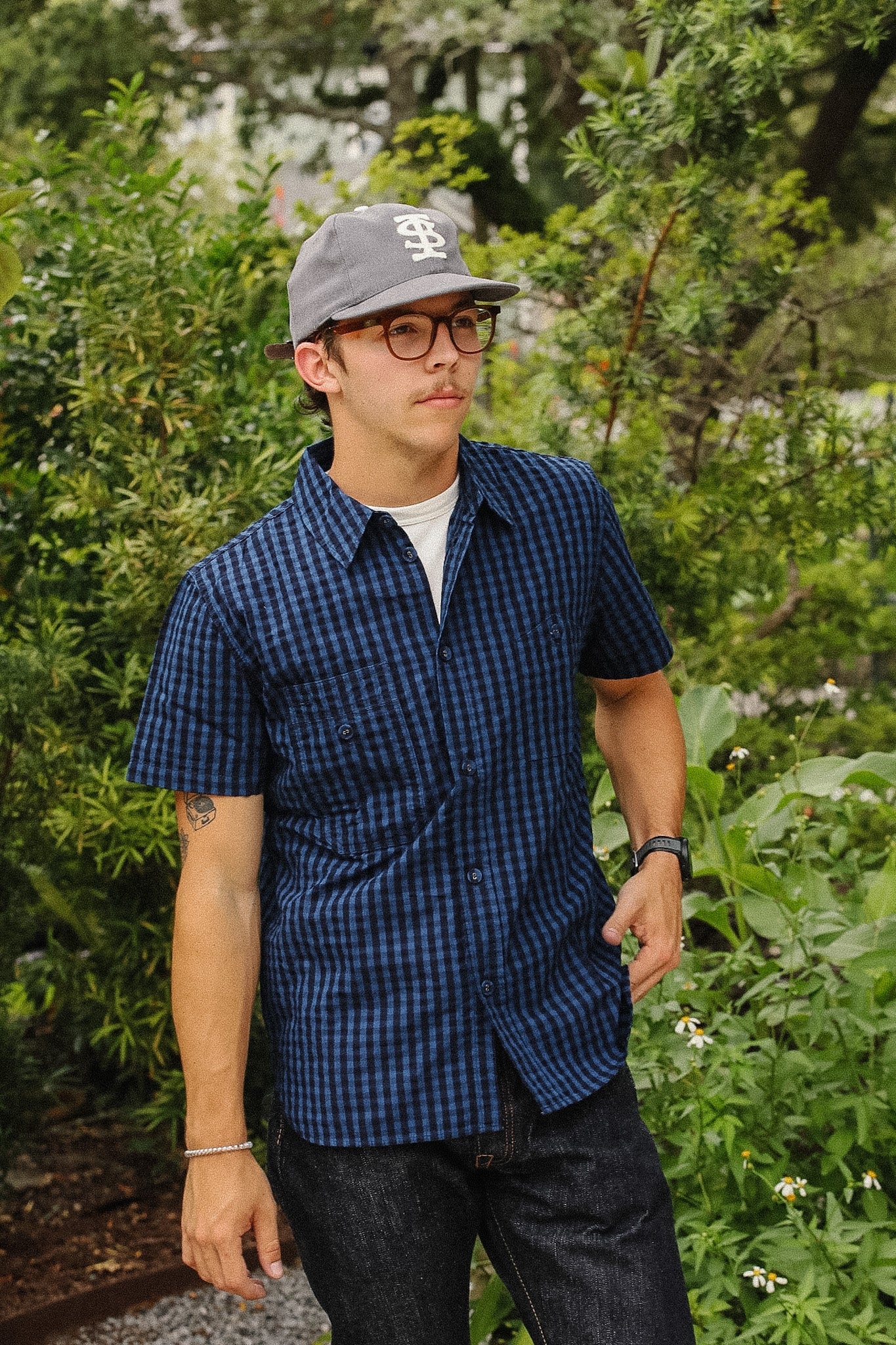 A man wearing a blue and black checkered short-sleeve button-up shirt, dark jeans, a gray baseball cap, and glasses, standing outdoors in a garden setting.