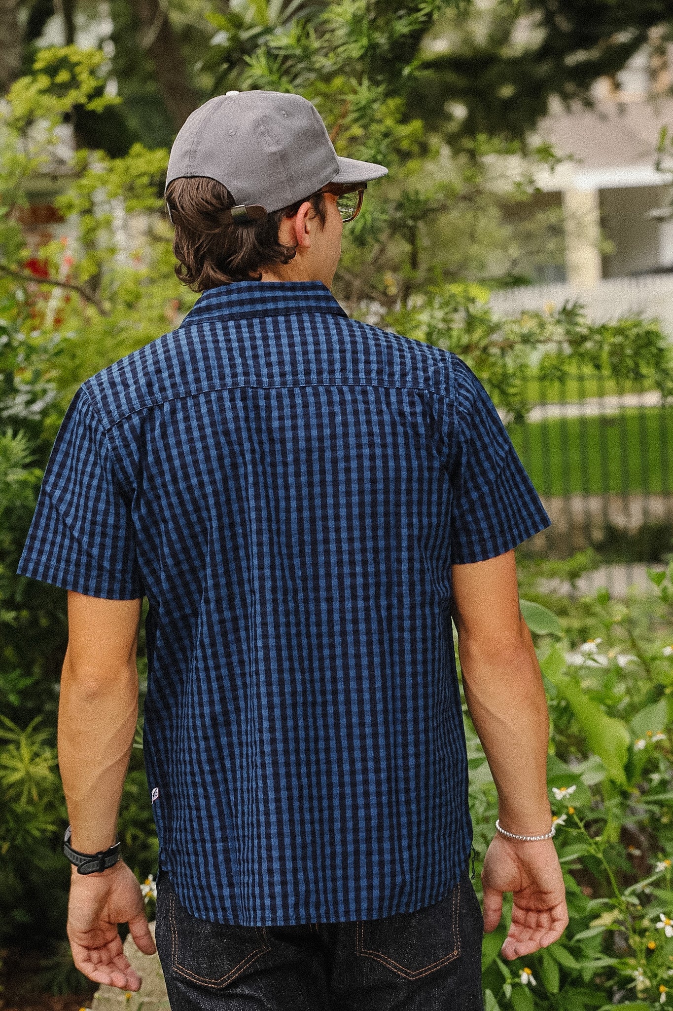 A man viewed from behind wearing a dark blue and black checkered short-sleeve button-up shirt, a gray baseball cap, and dark jeans, standing outdoors in a garden setting.