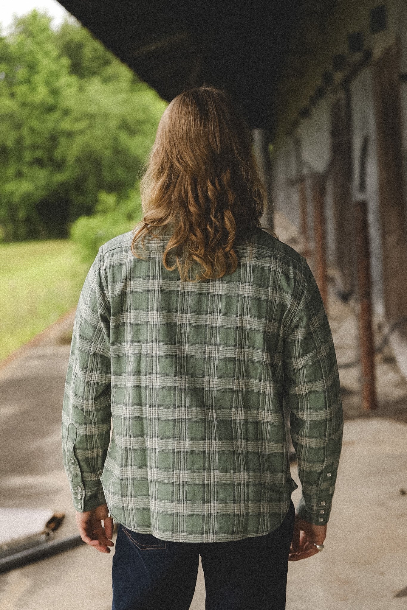 Person wearing a green plaid shirt standing outdoors with trees and a building in the background