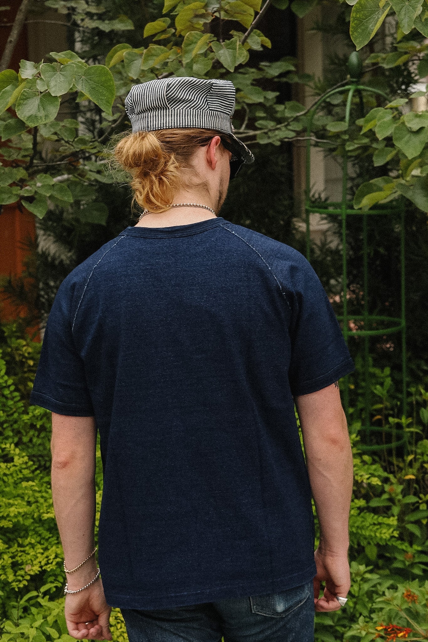 A person viewed from behind wearing a dark navy blue short-sleeve t-shirt with raglan sleeves, paired with a gray and white striped flat cap and jeans, standing outdoors in a garden setting.