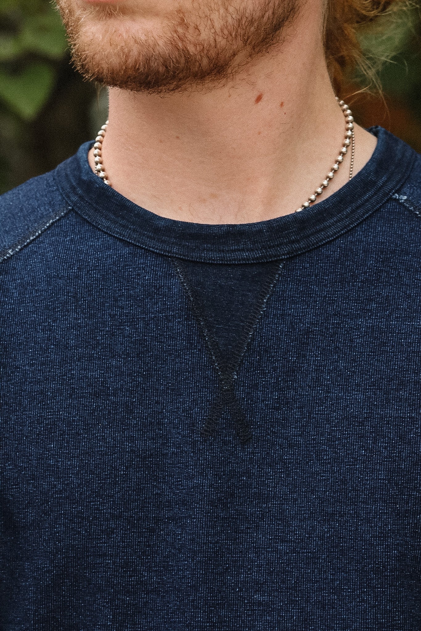 Close-up of a person wearing a dark navy crewneck sweatshirt and a silver beaded chain necklace, with a blurred outdoor background.