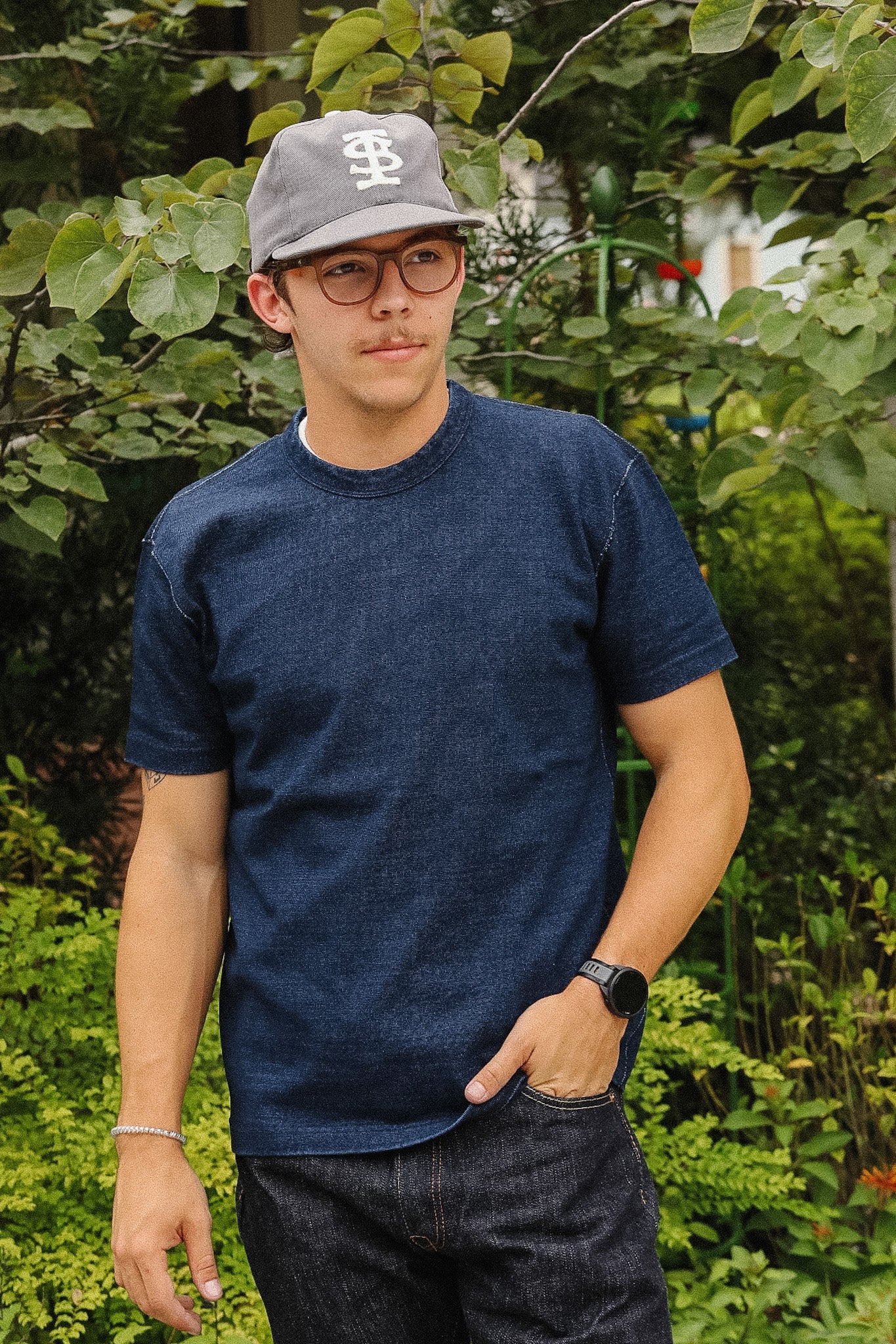A man viewed from behind wearing a dark blue short-sleeve t-shirt, a gray baseball cap, and dark jeans, standing outdoors in front of lush green foliage.