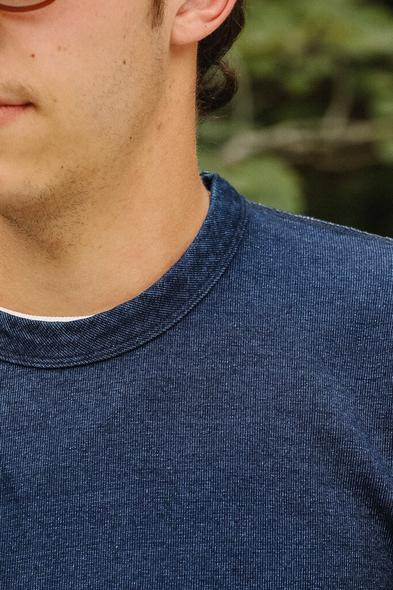 Close-up of a person wearing a dark navy blue crewneck knit shirt, showing the collar and textured fabric against a blurred green outdoor background.