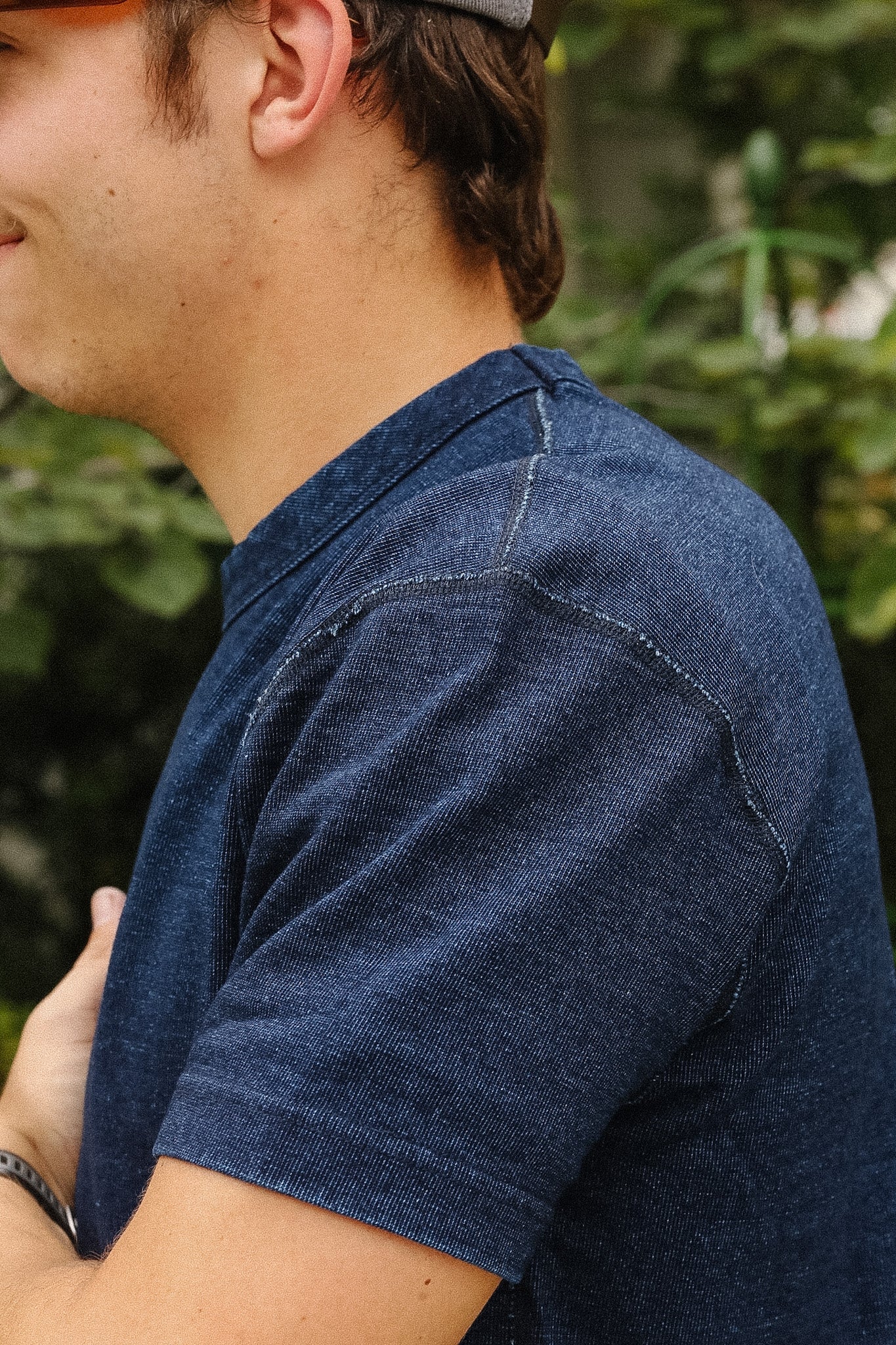 A close-up side profile of a man wearing a dark blue heathered short-sleeve t-shirt with visible contrast stitching, set against a blurred green outdoor background.