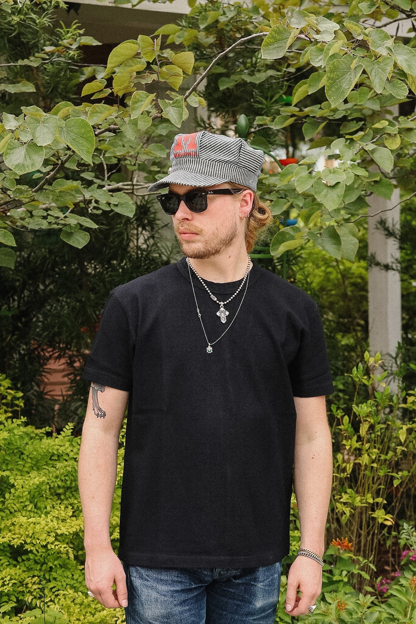 A man wearing a black crewneck t-shirt, blue jeans, a gray patterned cap, and sunglasses, standing outdoors in a garden setting.