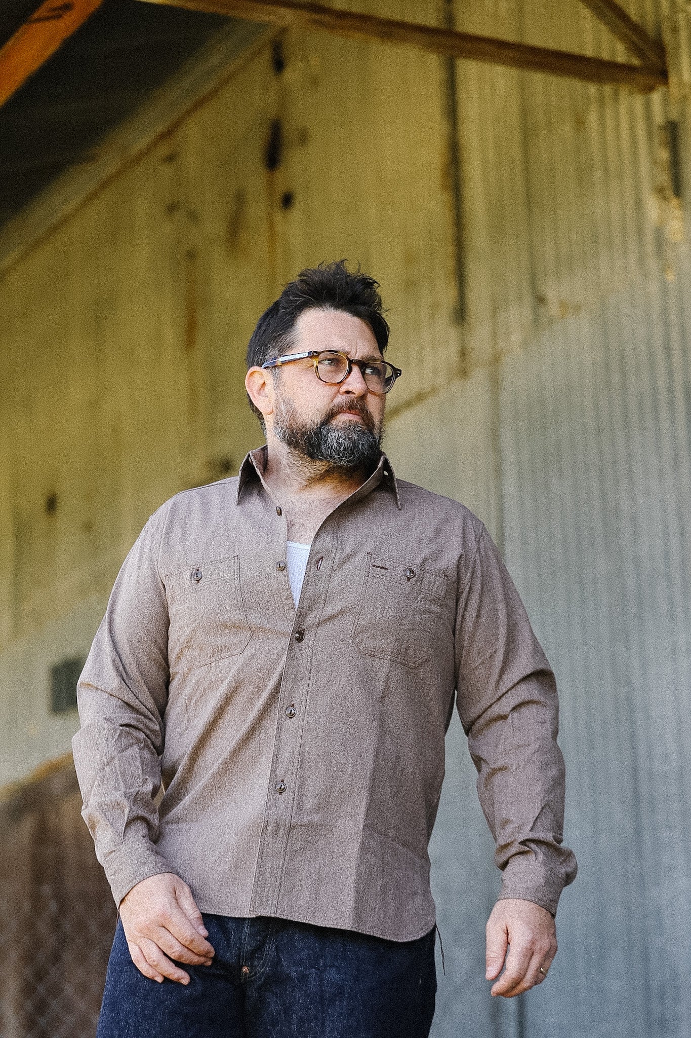 Man wearing glasses and a brown shirt standing against a rustic wall.
