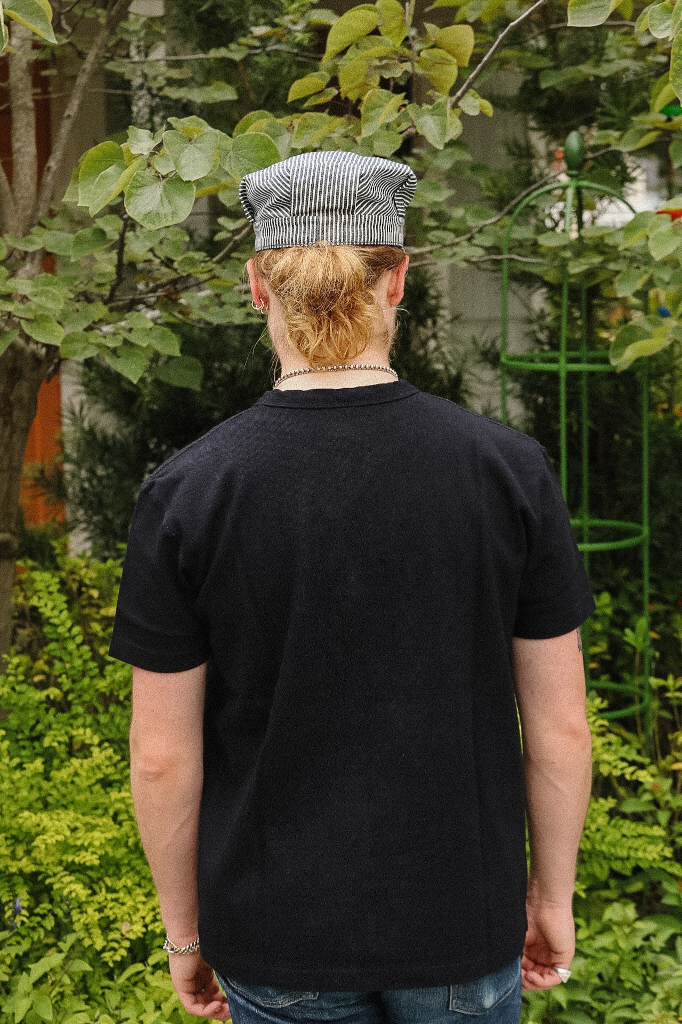 A man viewed from behind wearing a black crewneck t-shirt and a gray and white striped flat cap, standing outdoors in a garden setting.