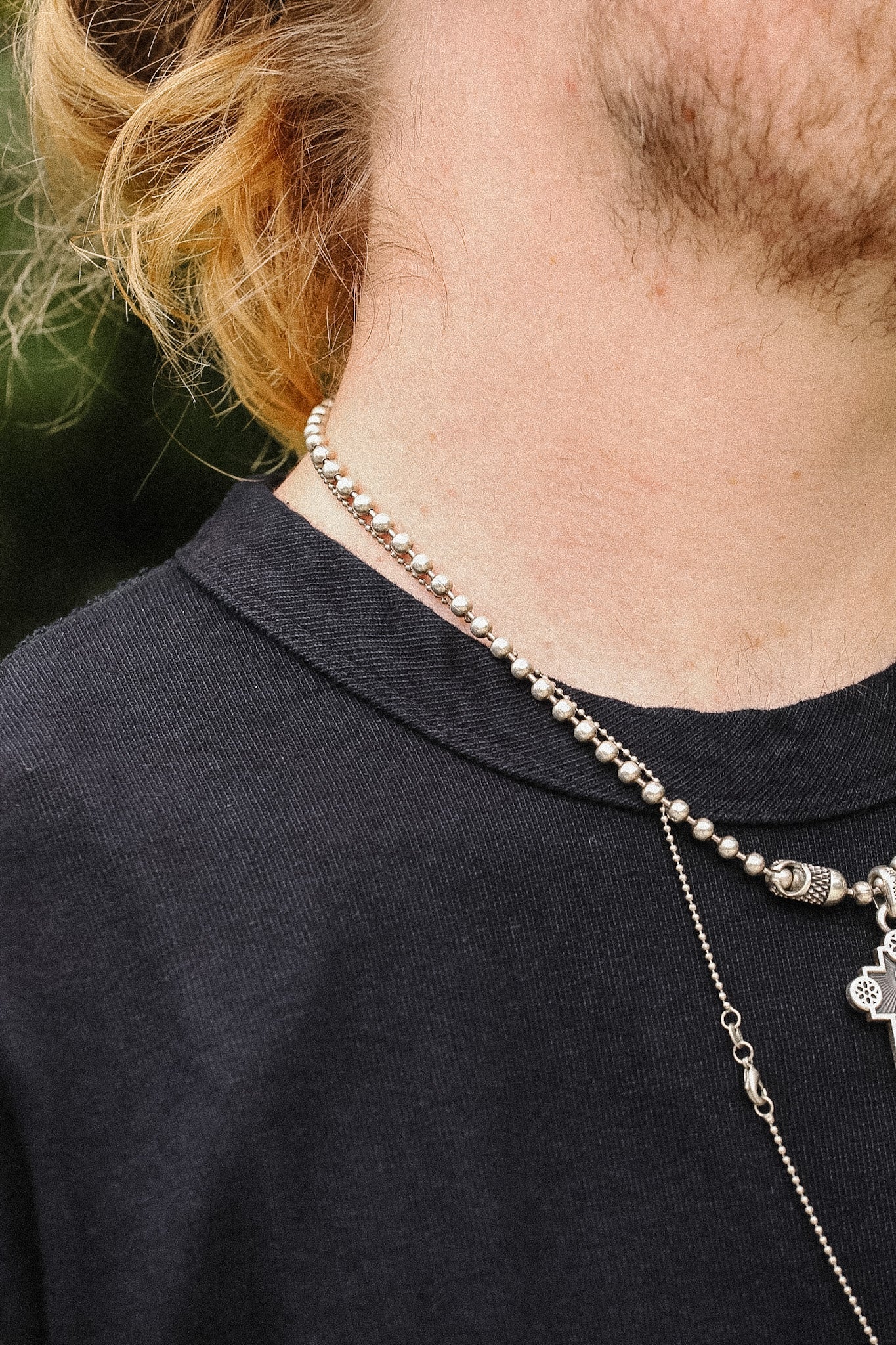 A close-up of a person's neck and upper chest wearing layered silver necklaces, including a beaded chain and a longer chain with a small skull pendant, over a black t-shirt.