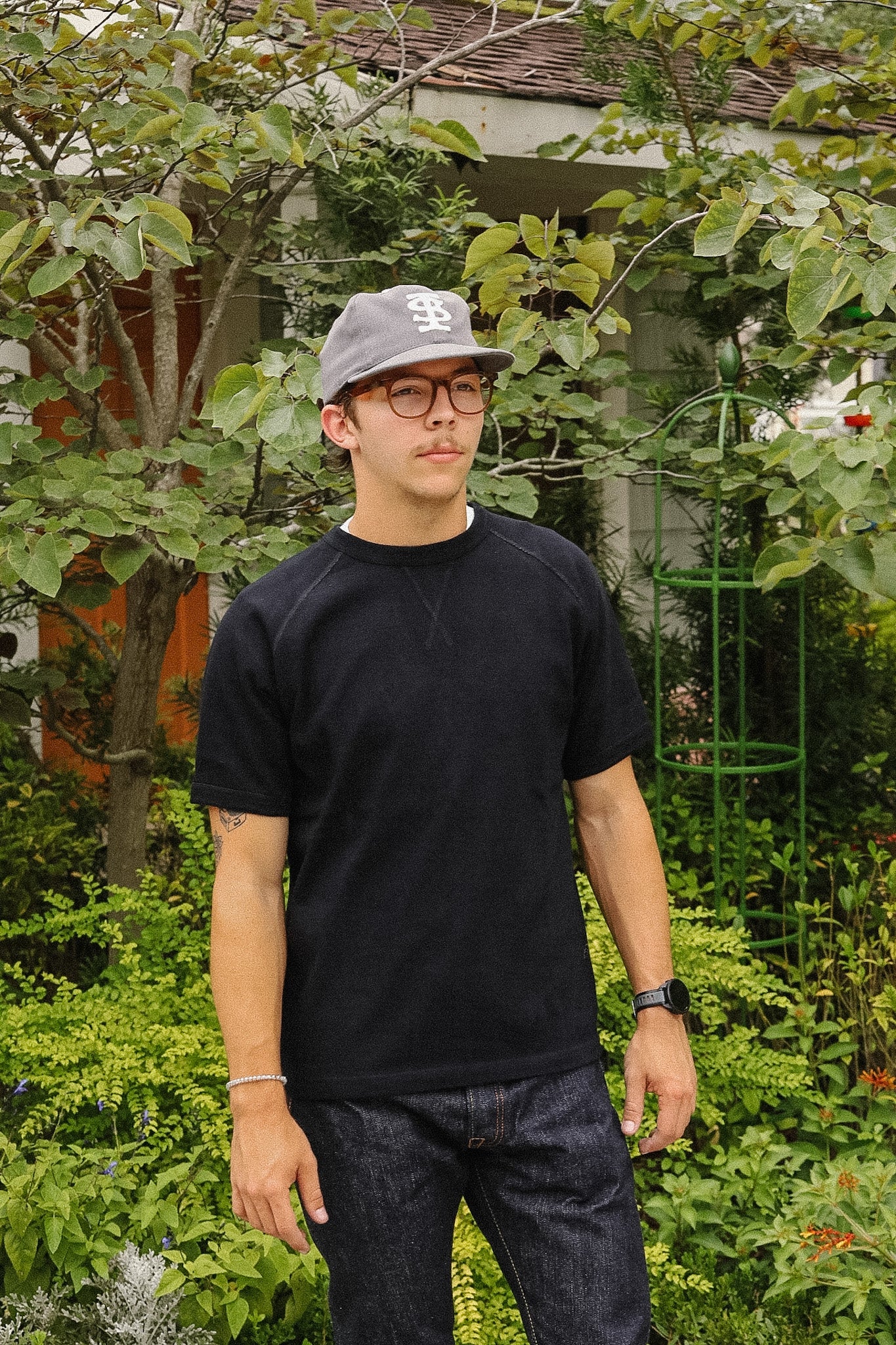 A man wearing a black crewneck t-shirt, dark jeans, a gray baseball cap, and glasses, standing in a lush green garden setting.