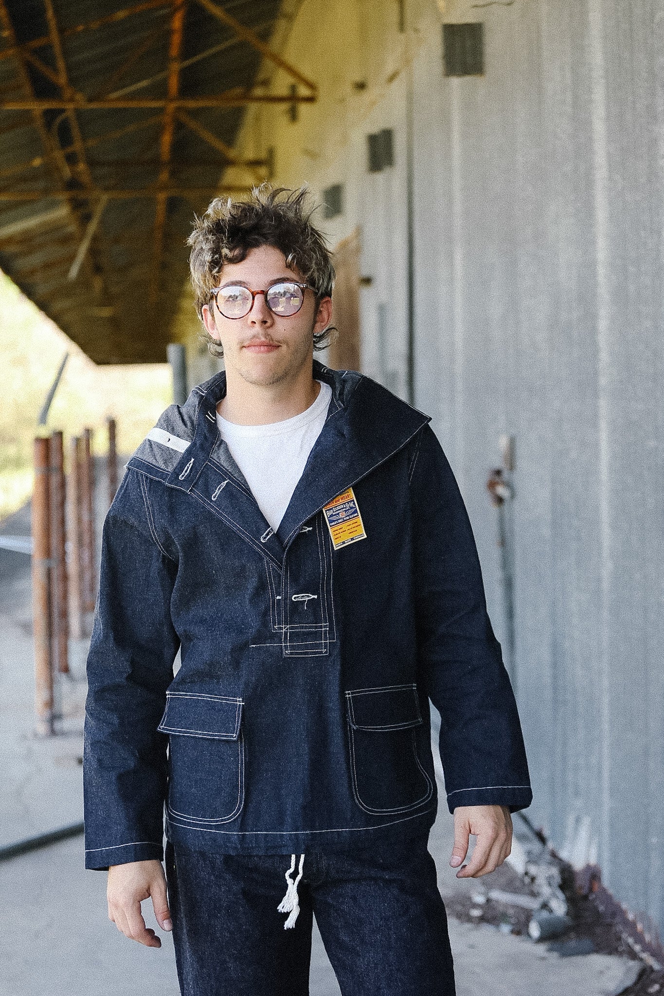 Person wearing a dark denim jacket and glasses standing in an industrial setting.