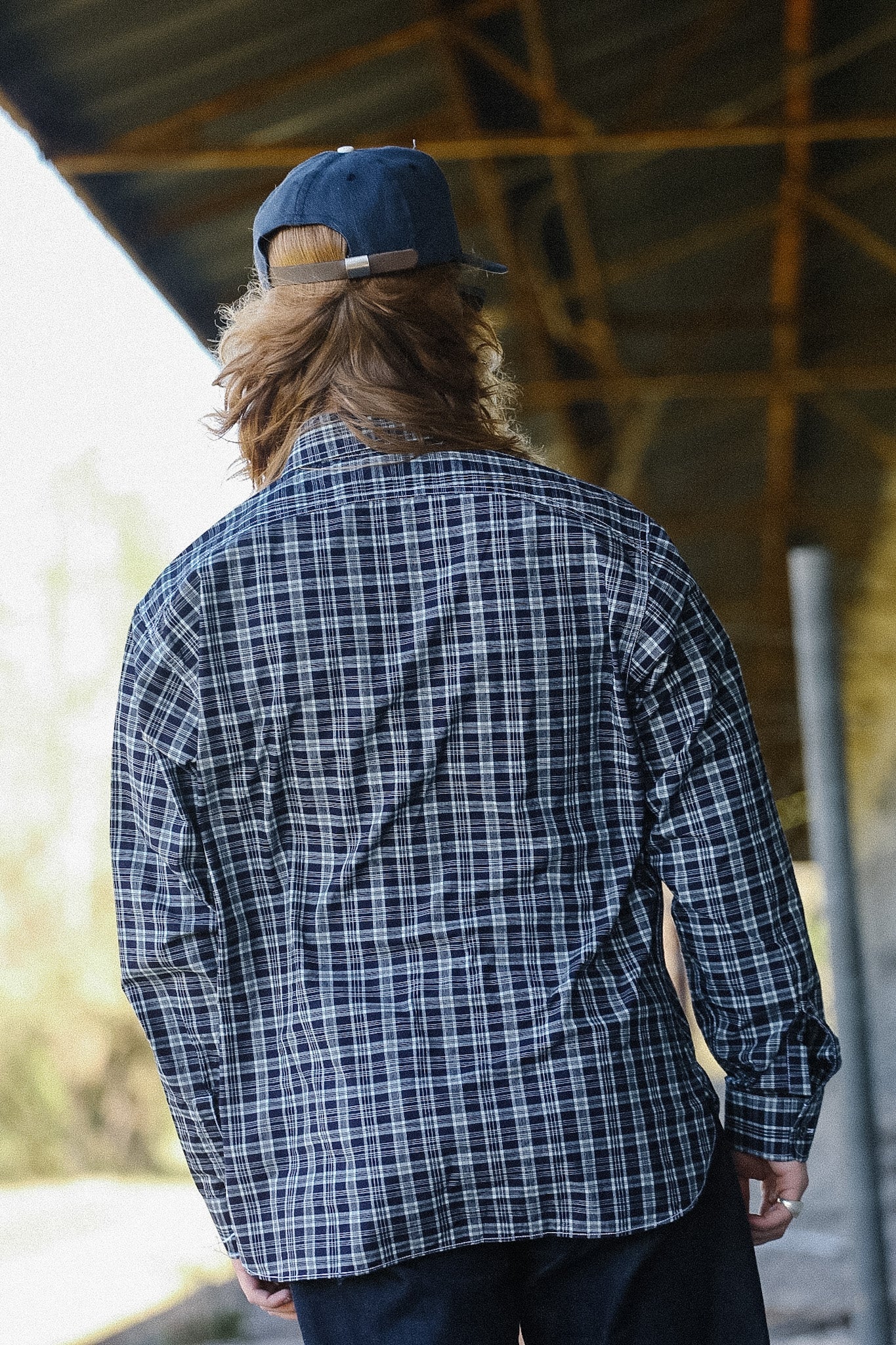 Person wearing a blue plaid shirt and cap from behind, standing under a wooden structure.
