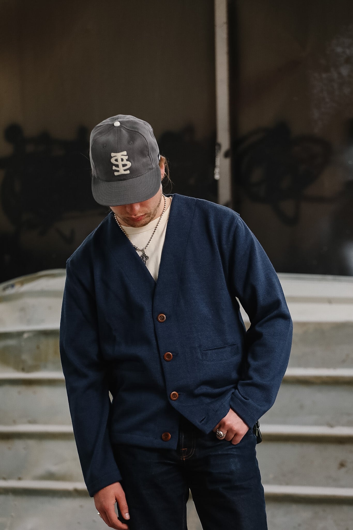 Person wearing a navy blue jacket with brown buttons and a gray cap, standing against a graffiti-covered wall.