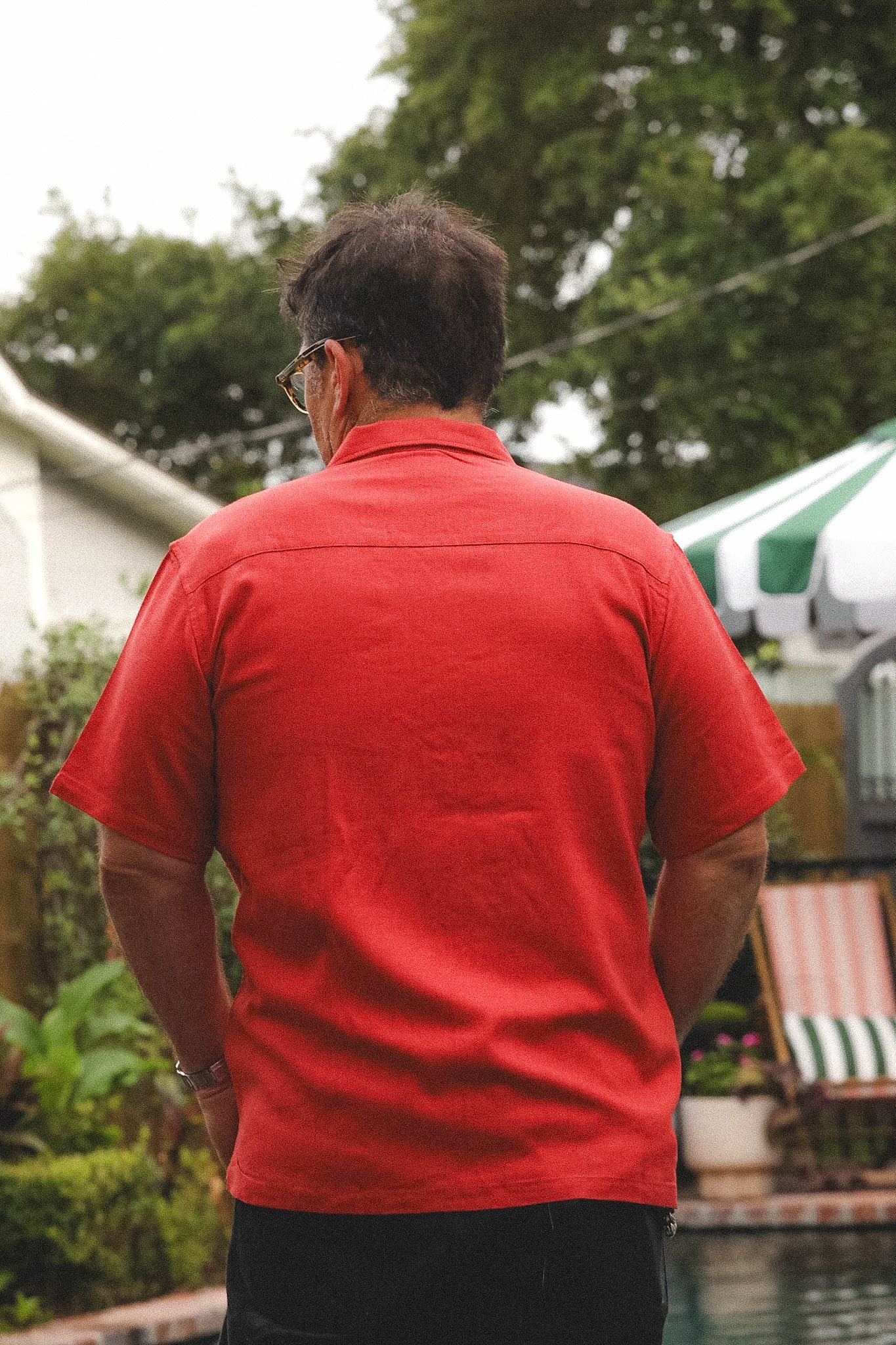 A man viewed from behind wearing a bright red short-sleeve button-up shirt and dark pants, standing outdoors near a pool with trees and patio furniture in the background.