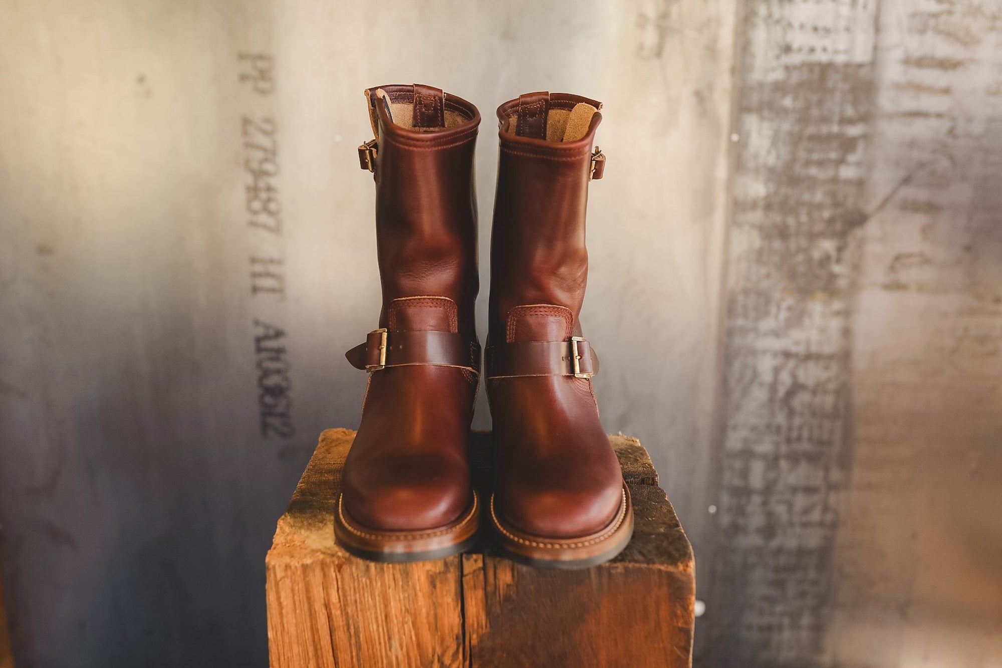 A pair of dark brown leather mid-calf boots with buckle straps, displayed on a rustic wooden block against a textured, warm-toned industrial background.