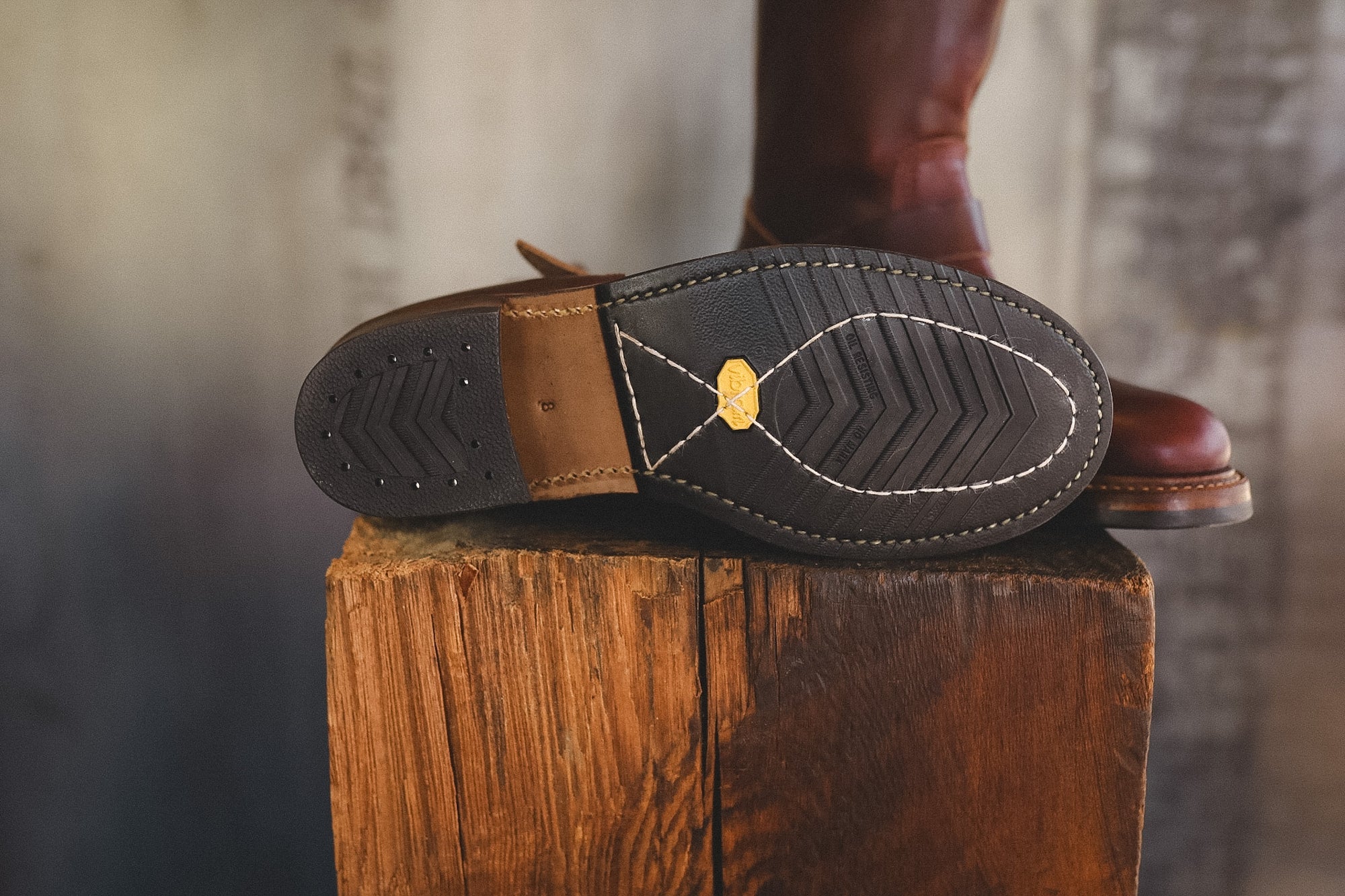 A close-up of a brown leather boot's sole resting on a rustic wooden block, with the boot itself partially visible in the background against a blurred industrial-style backdrop.