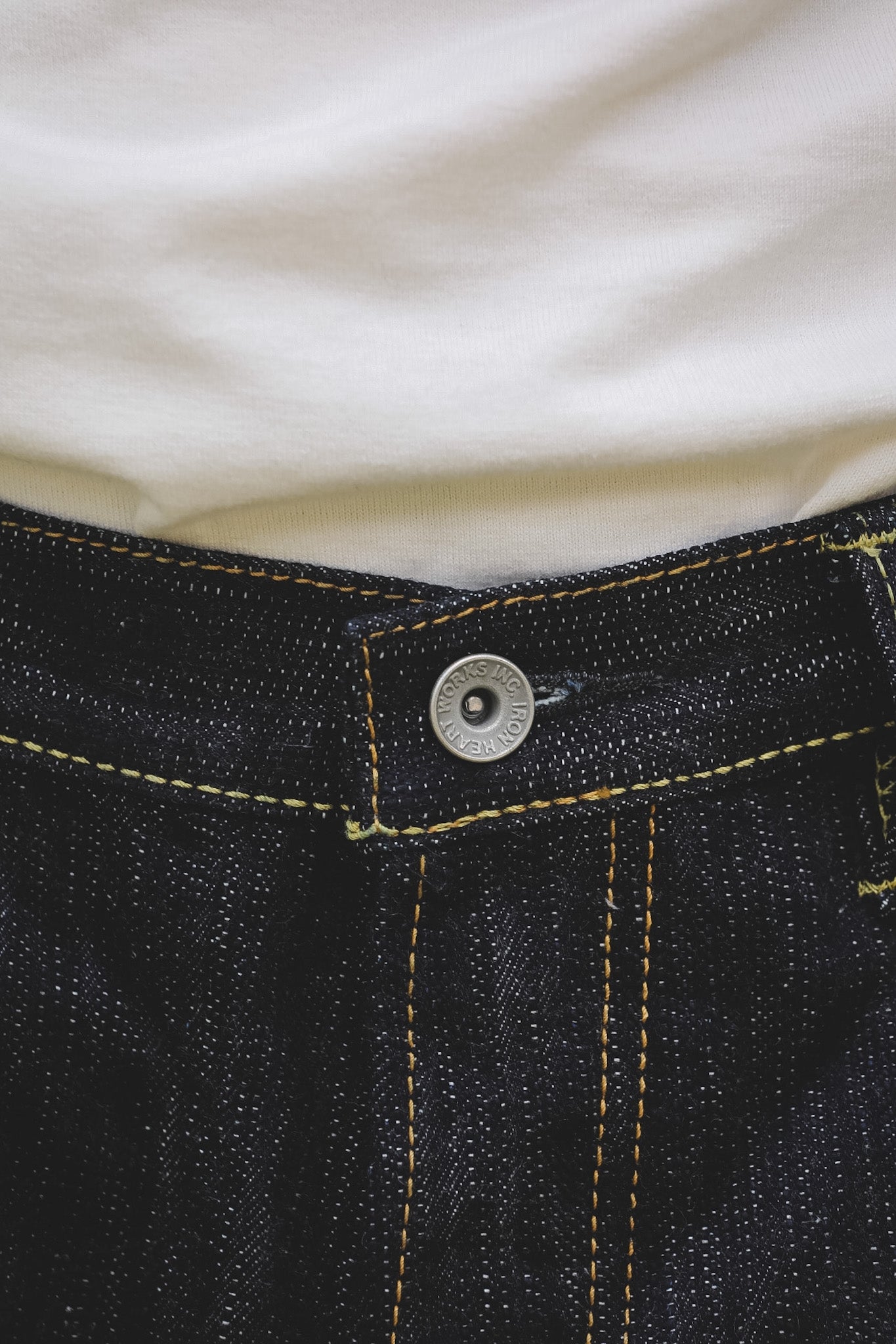 Close-up of dark indigo denim jeans with yellow contrast stitching, a silver metal button, and a white t-shirt tucked in.