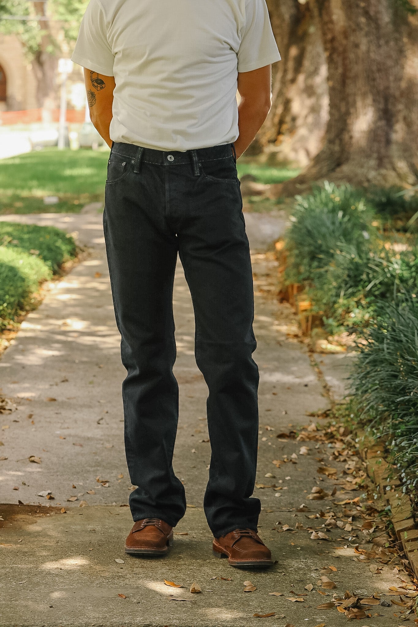 A person standing on a tree-lined path wearing dark charcoal gray jeans, a white t-shirt, and brown shoes.