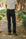 A person standing on a tree-lined path wearing dark charcoal gray jeans, a white t-shirt, and brown shoes.