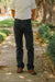 A person standing on a tree-lined path wearing dark charcoal gray jeans, a white t-shirt, and brown shoes.