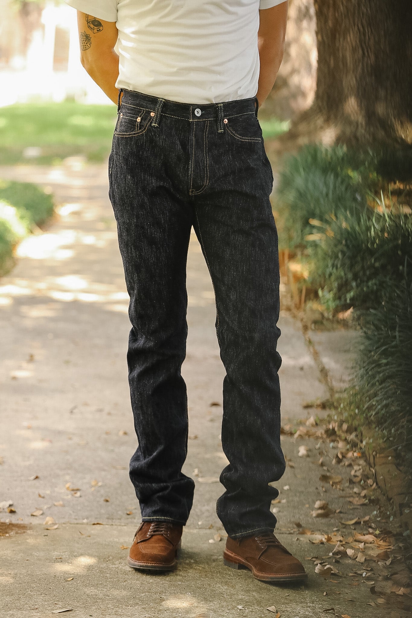 A person standing outdoors on a paved path wearing dark indigo denim jeans, a white t-shirt, and brown leather shoes.