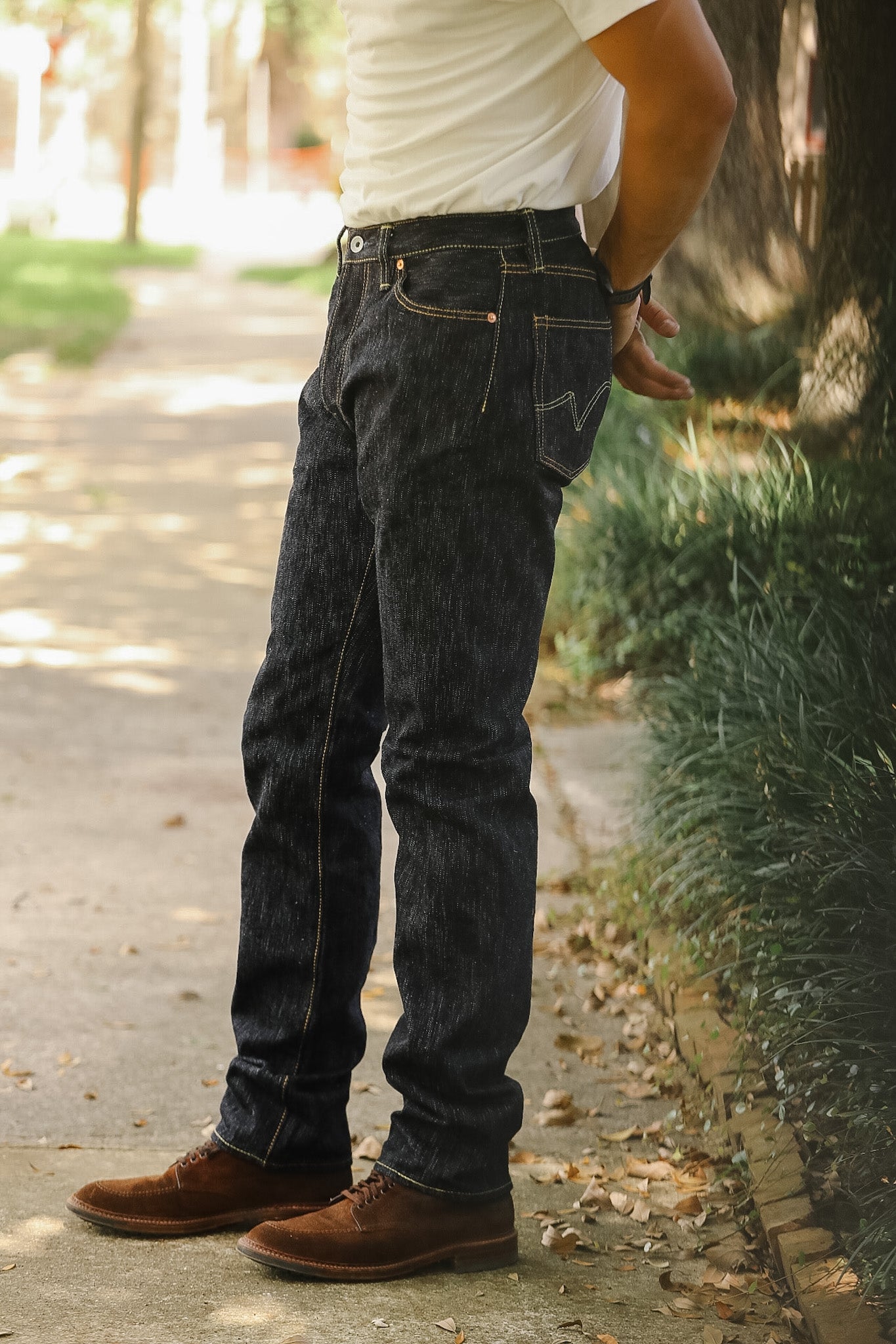 A person standing outdoors on a paved path wearing dark indigo denim jeans, a white t-shirt, and brown leather shoes.