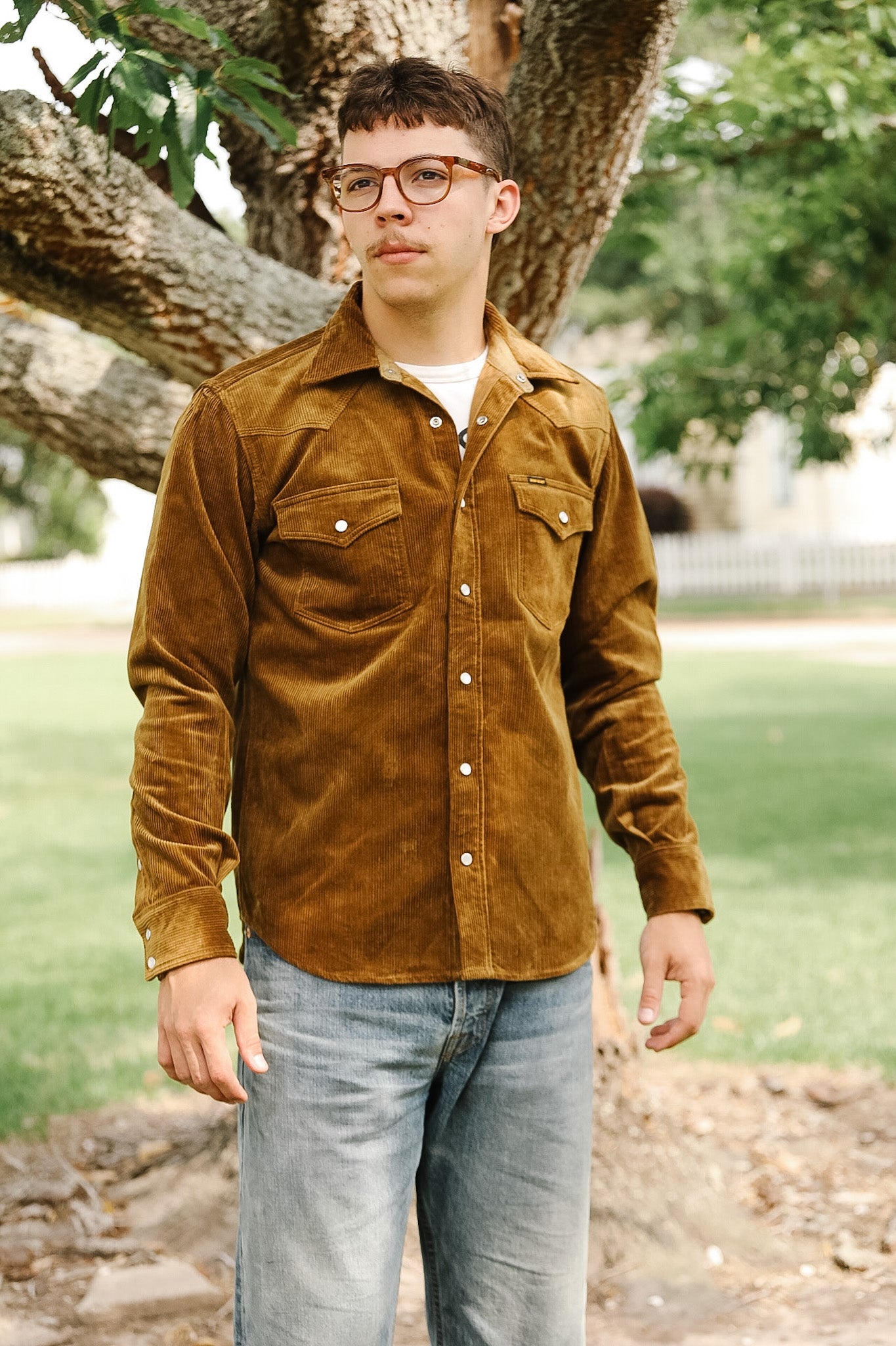A man wearing a brown corduroy western shirt with a collar, button-up front, and cap, standing in front of a white background with a ladder in the background.
