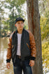 Young man wearing Iron Shop cap and leather-sleeve jacket standing by tree near lake in outdoor setting