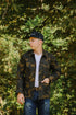 Young man in camo jacket and black Iron Shop cap standing outdoors with green tree background